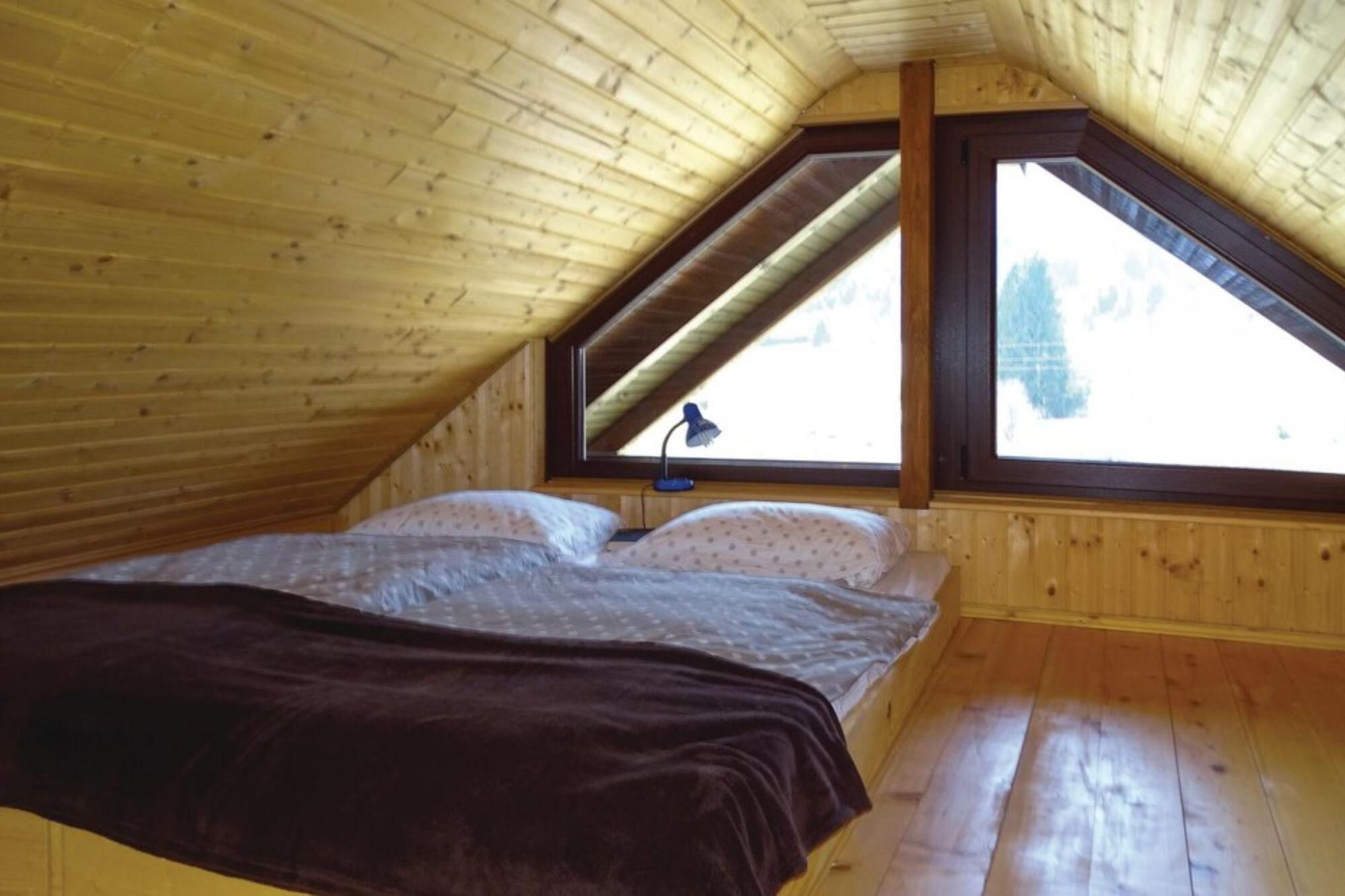 A bedroom with a wooden sloped ceiling, a bed with a dark blanket, and a window with a view of trees.