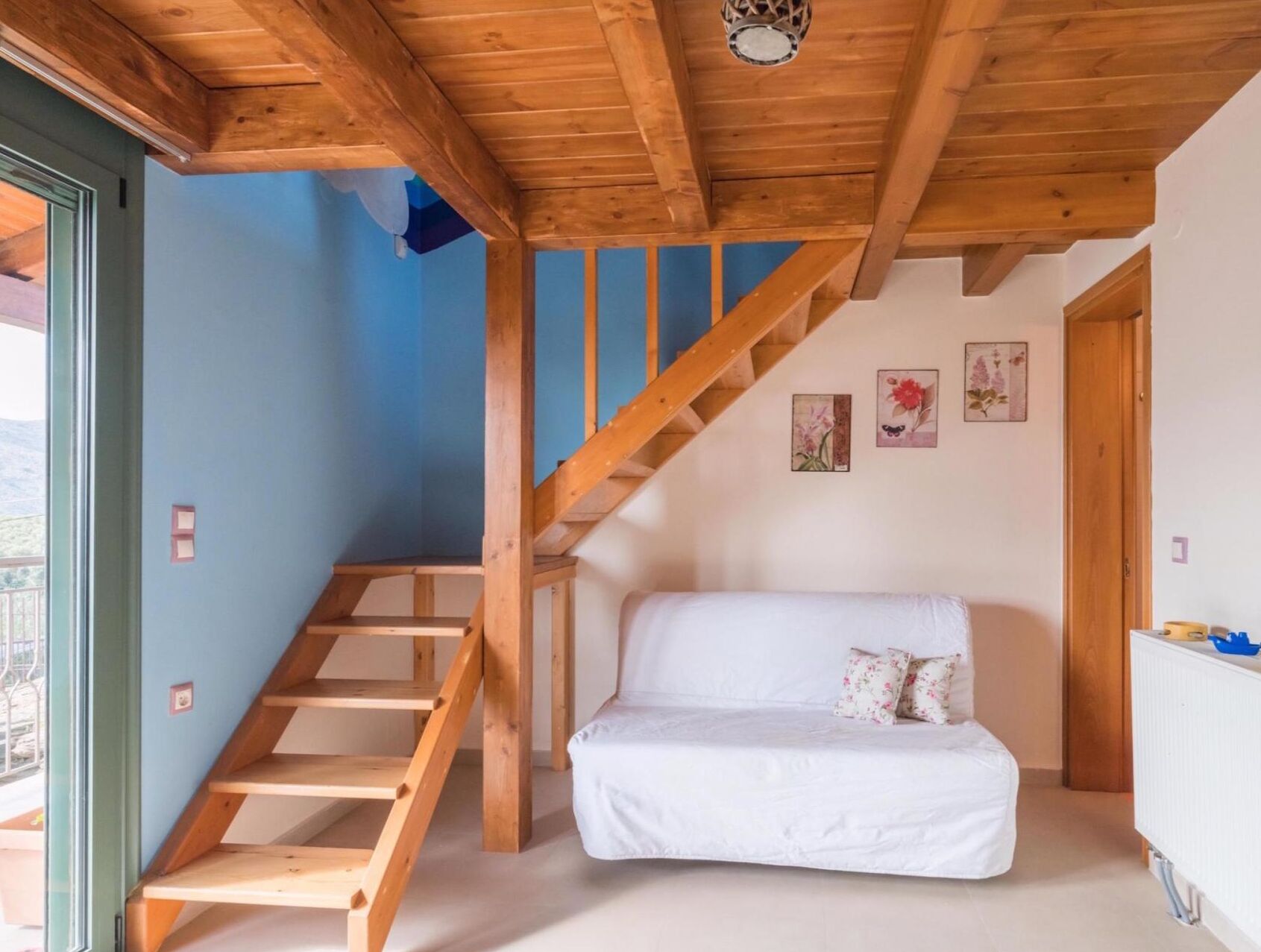 A room with a wooden staircase, a white sofa, and a view through a glass door.