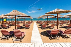 A beach with rows of striped beach chairs and thatched umbrellas, a wooden walkway, and a clear blue sky. - Villa Eolos Crete in Heraklion (Malevizi)