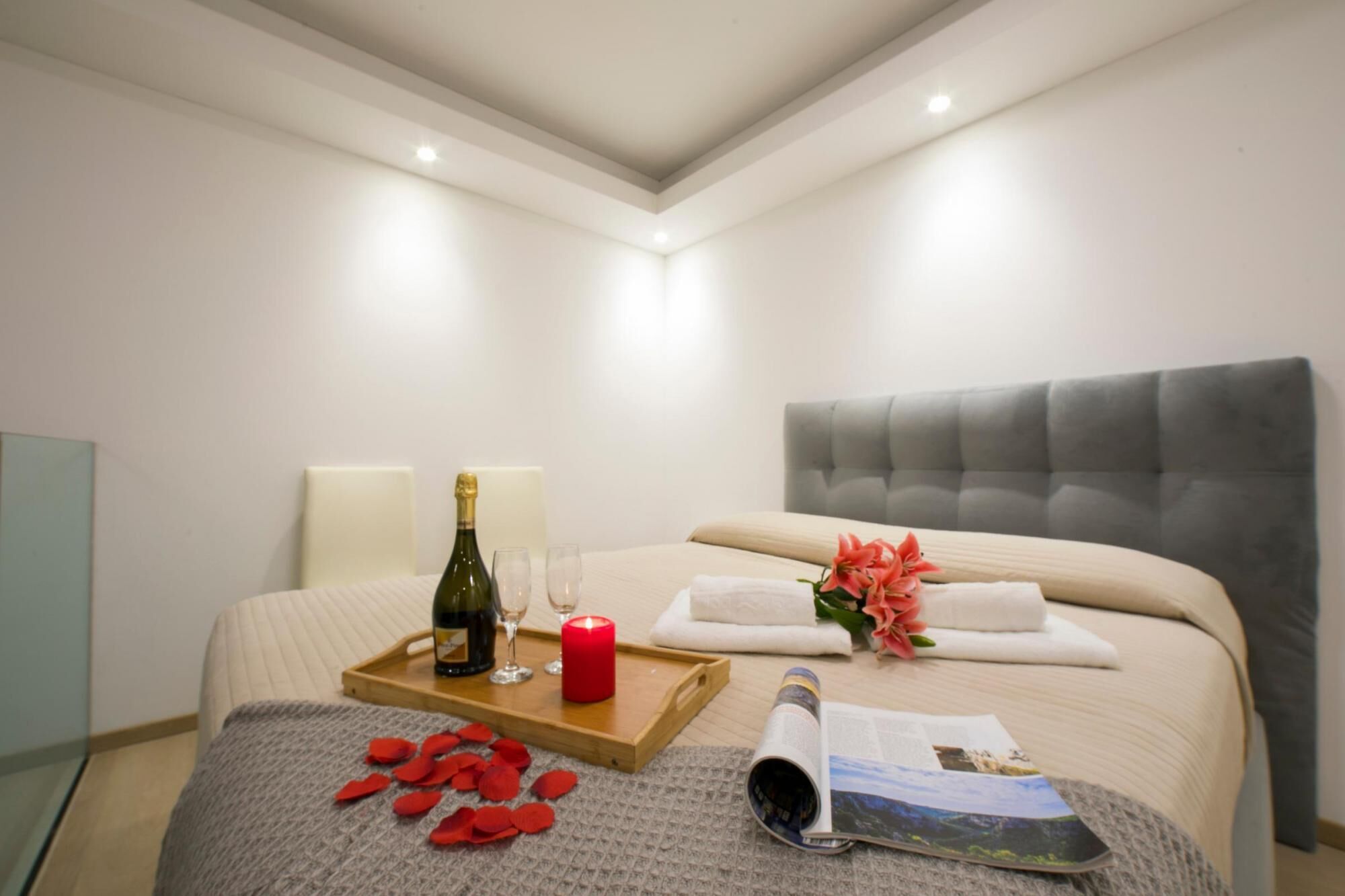 Une chambre d’hôtel avec un lit, une table de chevet sur laquelle se trouve une bouteille de champagne, des verres, une bougie rouge, un plateau parsemé de pétales de rose et un magazine.