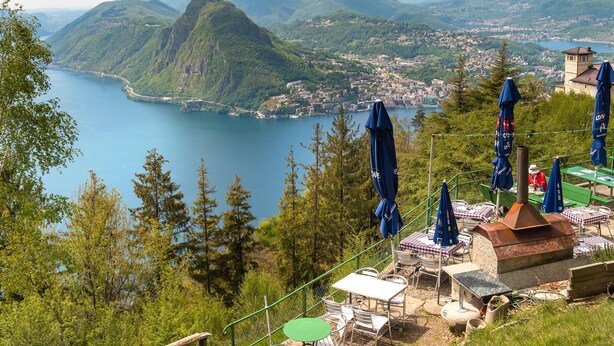 A lakeside terrace with outdoor seating and a view of a town and mountains. - Rebecca Garden in Lugano (Lugano)