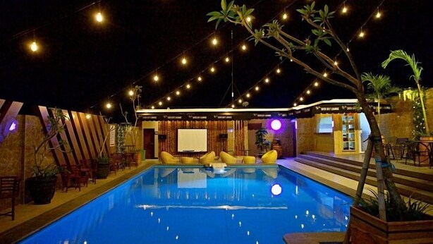 A swimming pool with string lights overhead, surrounded by wooden deck chairs and tables. - THE SATO HOTEL (Kudus)