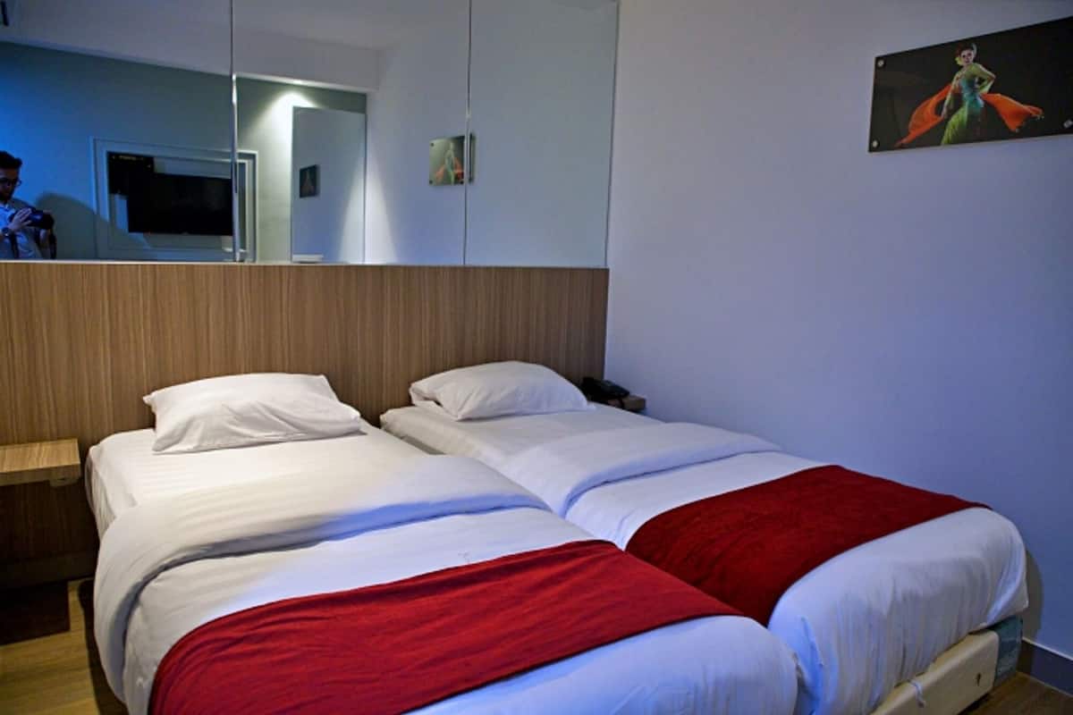 Habitación de hotel con dos camas, cabecera de madera, manta roja, un cuadro en la pared y un espejo que refleja parte de la habitación.