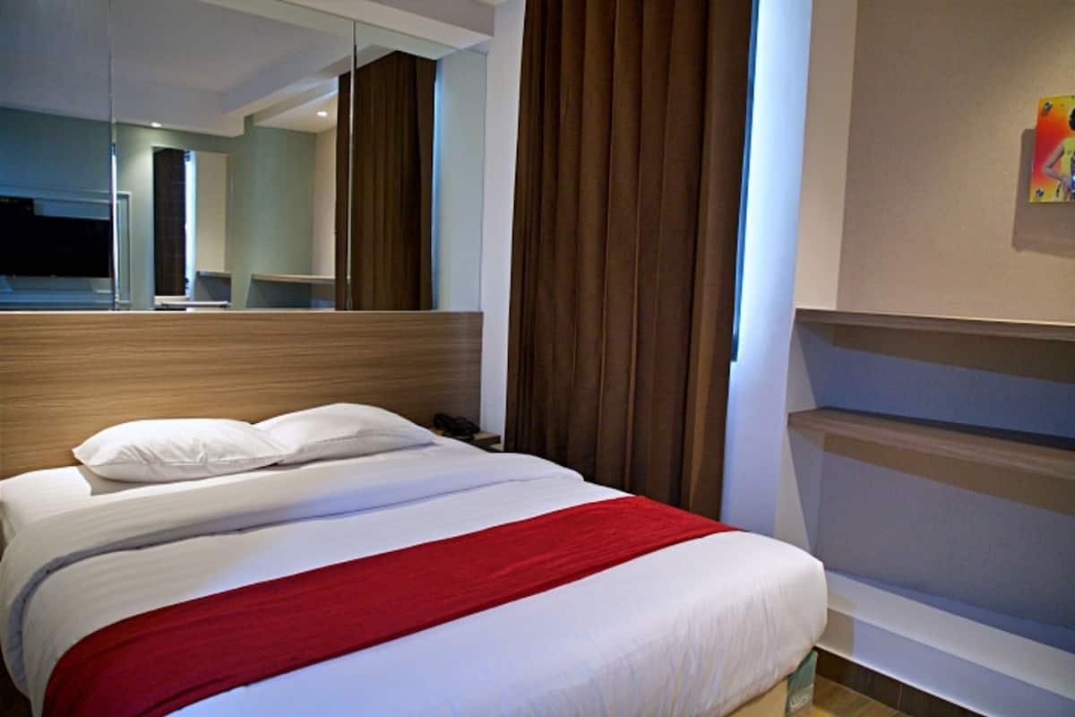 Habitación de hotel con una cama grande, cabecero de madera, manta roja y un espejo amplio, además de un ventanal con cortinas.