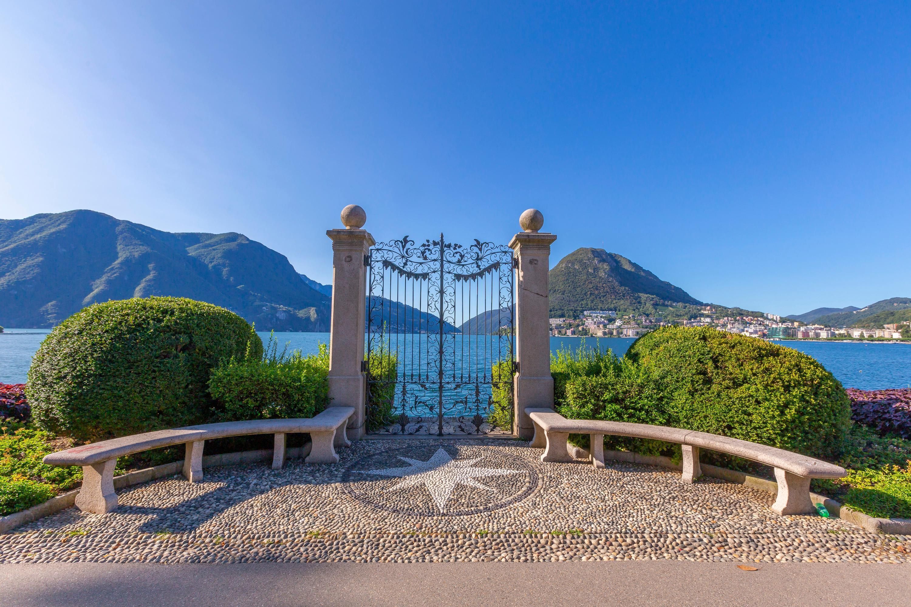 A stone bench and gate leading to a scenic view of a lake and mountains.