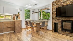 A modern kitchen with wooden cabinets, a dining area with a table and chairs, a large window with a view of greenery, and a fireplace with a TV mounted above it. - Pool Villa Katja Kranjska Gora (Kranjska Gora)