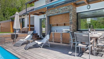 Un área de piscina moderna al aire libre con sillones de descanso, una parrilla y una terraza de madera.