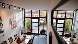 Un espace de restauration moderne avec une longue table, des chaises et des plantes en pot. De grandes fenêtres offrent une vue sur l’extérieur.