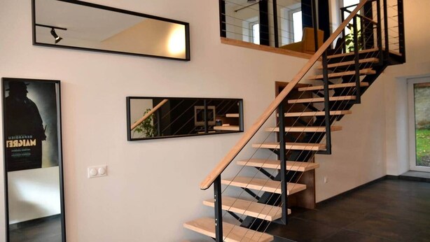 Un escalier moderne avec des marches en bois et des rampes noires, flanqué de deux miroirs fixés au mur et d’un tableau encadré.