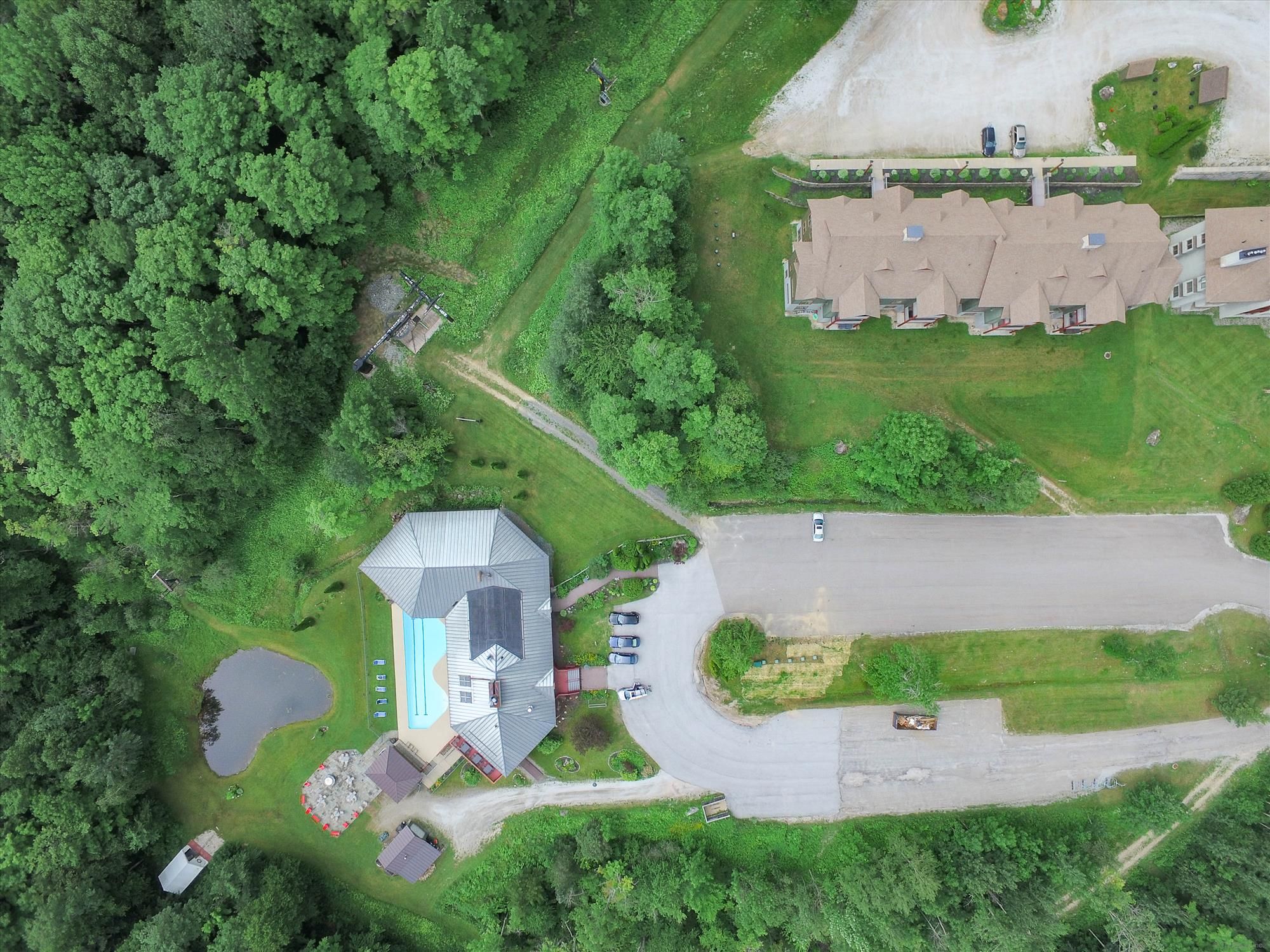 An aerial view of a residential area with a large house featuring a swimming pool, multiple cars parked, and a well-maintained lawn.