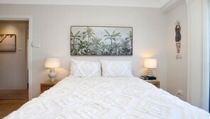 A neatly arranged bedroom with a large bed, two bedside lamps, a tropical-themed painting, and a decorative wall piece. - Seacrest 7 - Beach Access Across the Road (Hawks Nest)