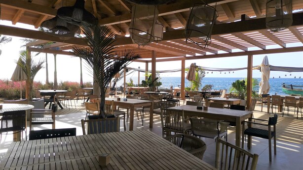 Un espace de restauration en plein air avec des tables et des chaises en bois, des lampes suspendues et une vue sur la mer.