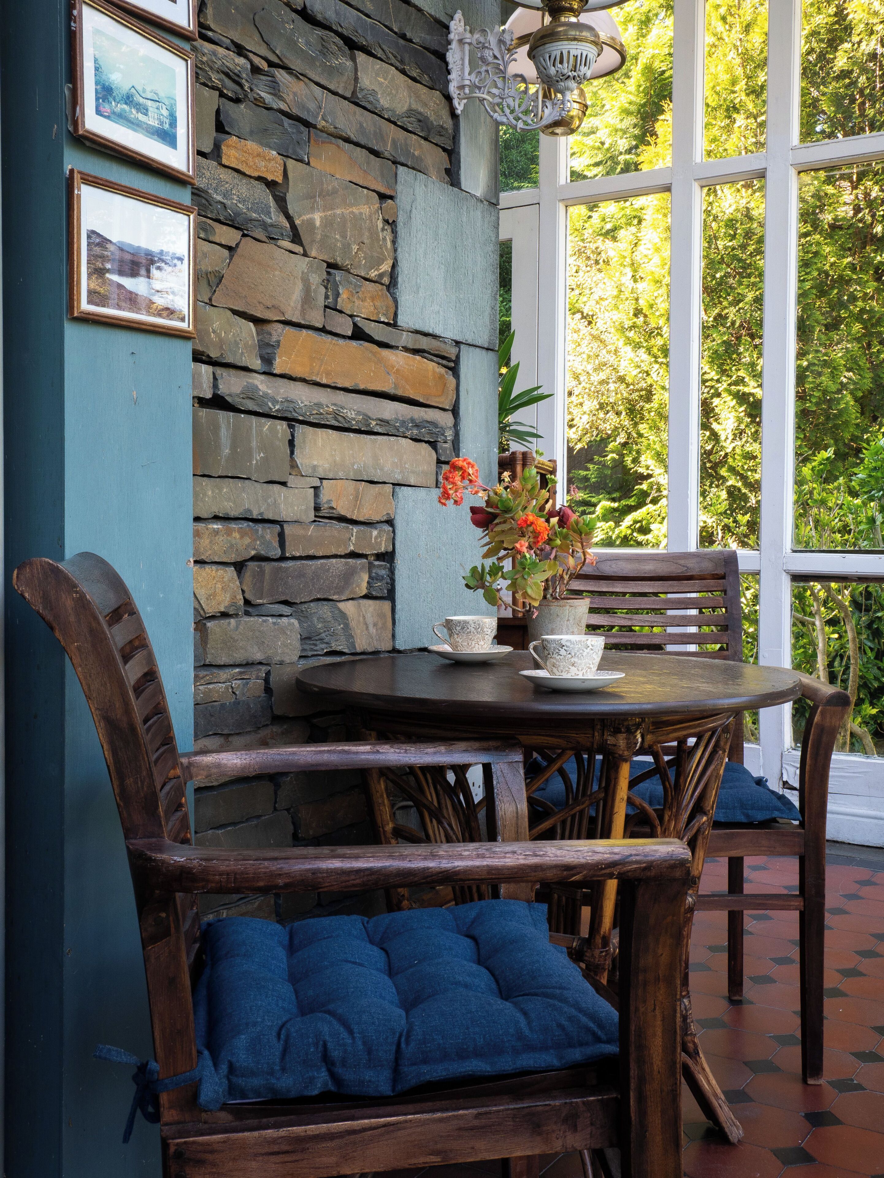 A wooden dining table with chairs, a stone wall, and a window with a view of greenery.