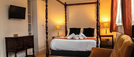 A four-poster bed with a wooden frame, a television mounted on the wall, a wooden nightstand with a lamp, and a leather armchair.
