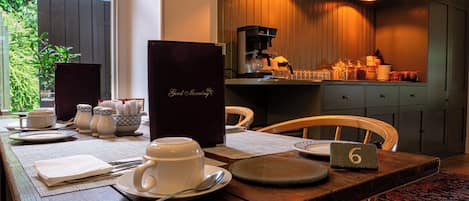 Una mesa de desayuno dispuesta con tazas de té, platos y una cafetera, en una cocina acogedora con armarios de madera y vistas a zonas verdes.