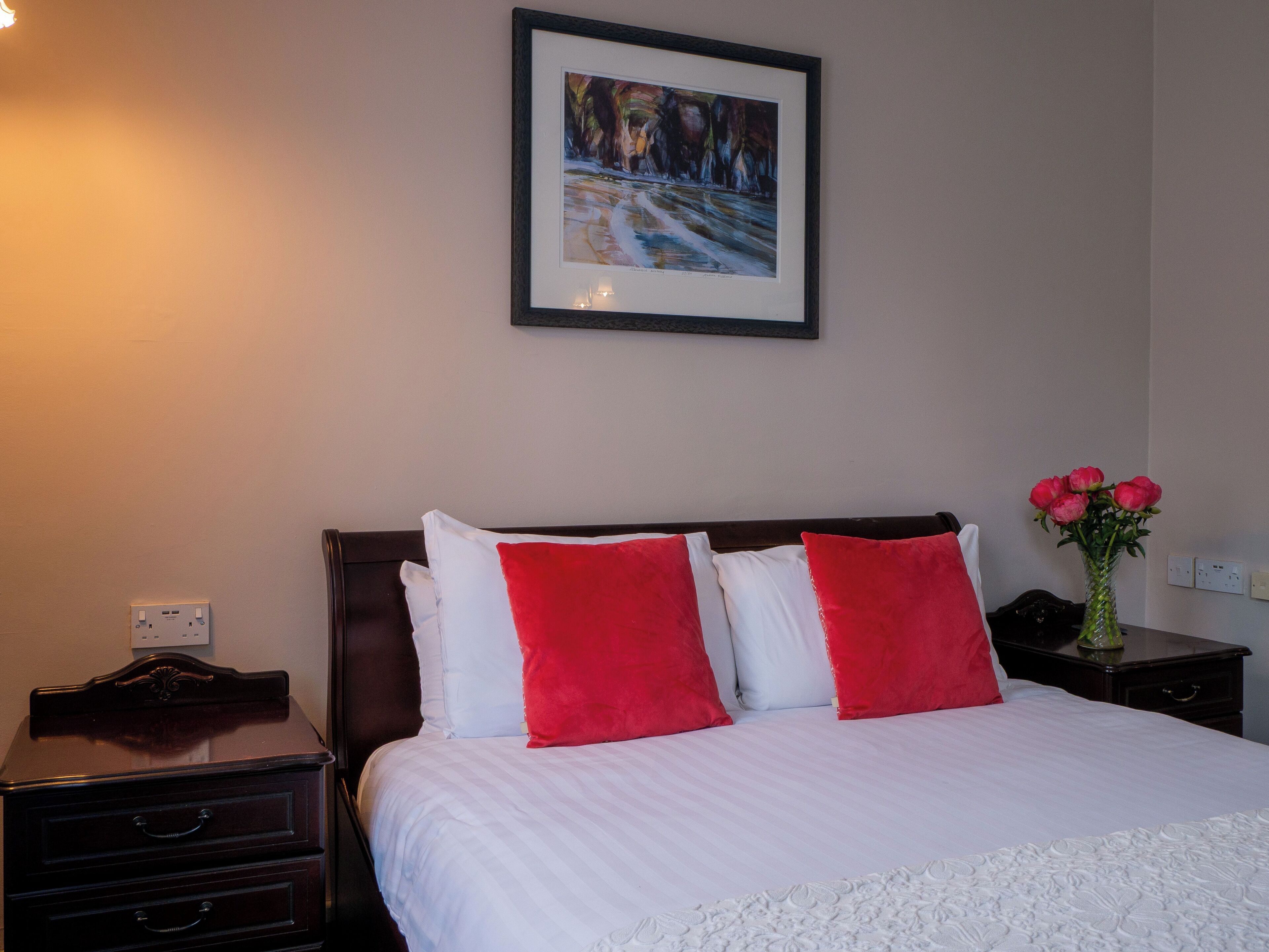 Una camera d'albergo con un letto, un comodino, un vaso con fiori e un quadro appeso al muro.