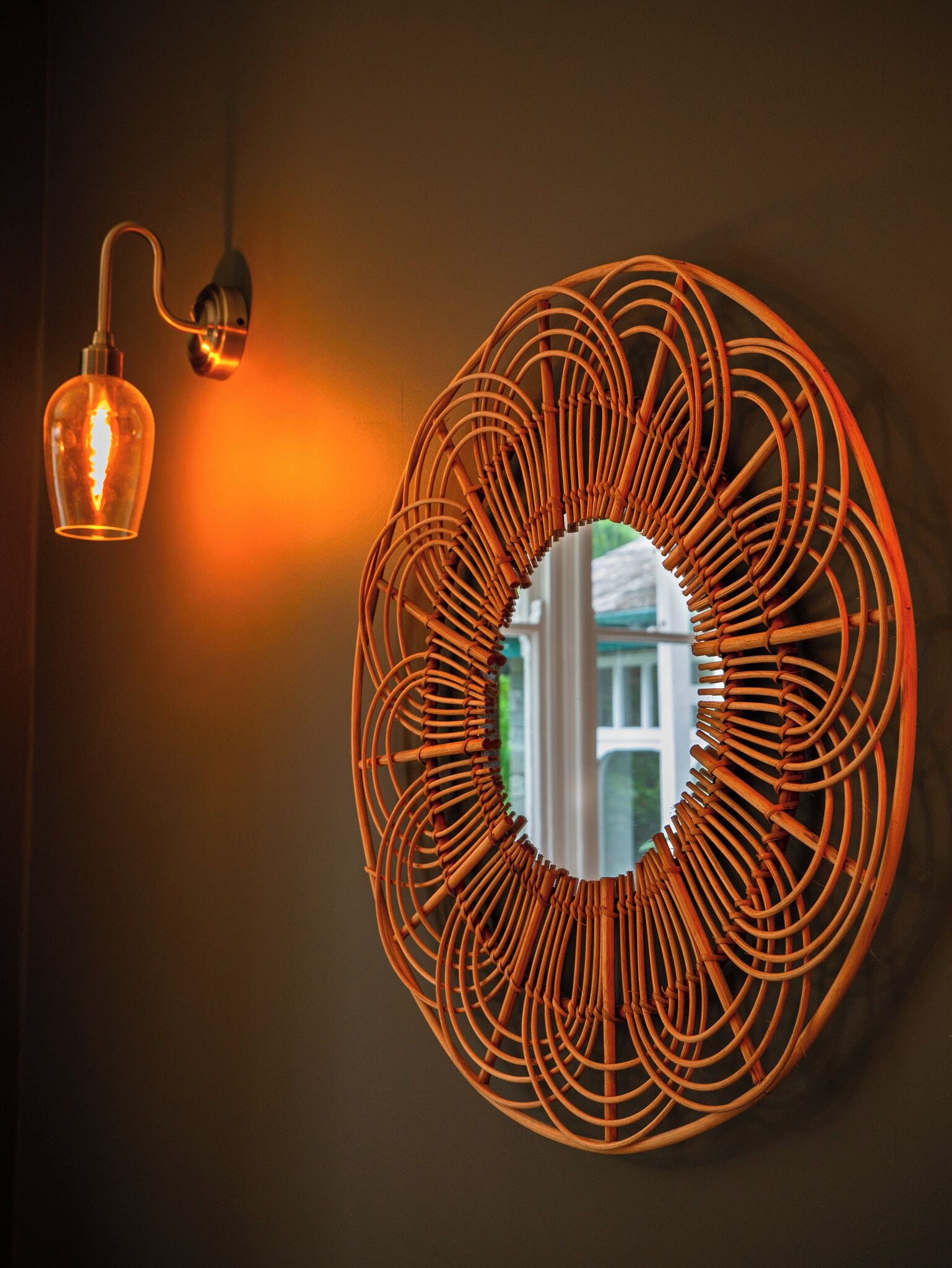 Uno specchio circolare fissato al muro, con cornice intrecciata, un lampadario a sospensione e il riflesso di un edificio.