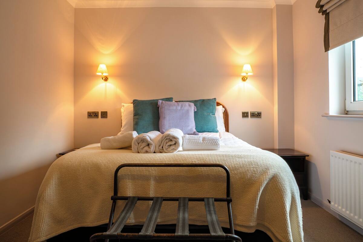 A neatly made bed with a headboard, two wall-mounted lamps, and a window with curtains.