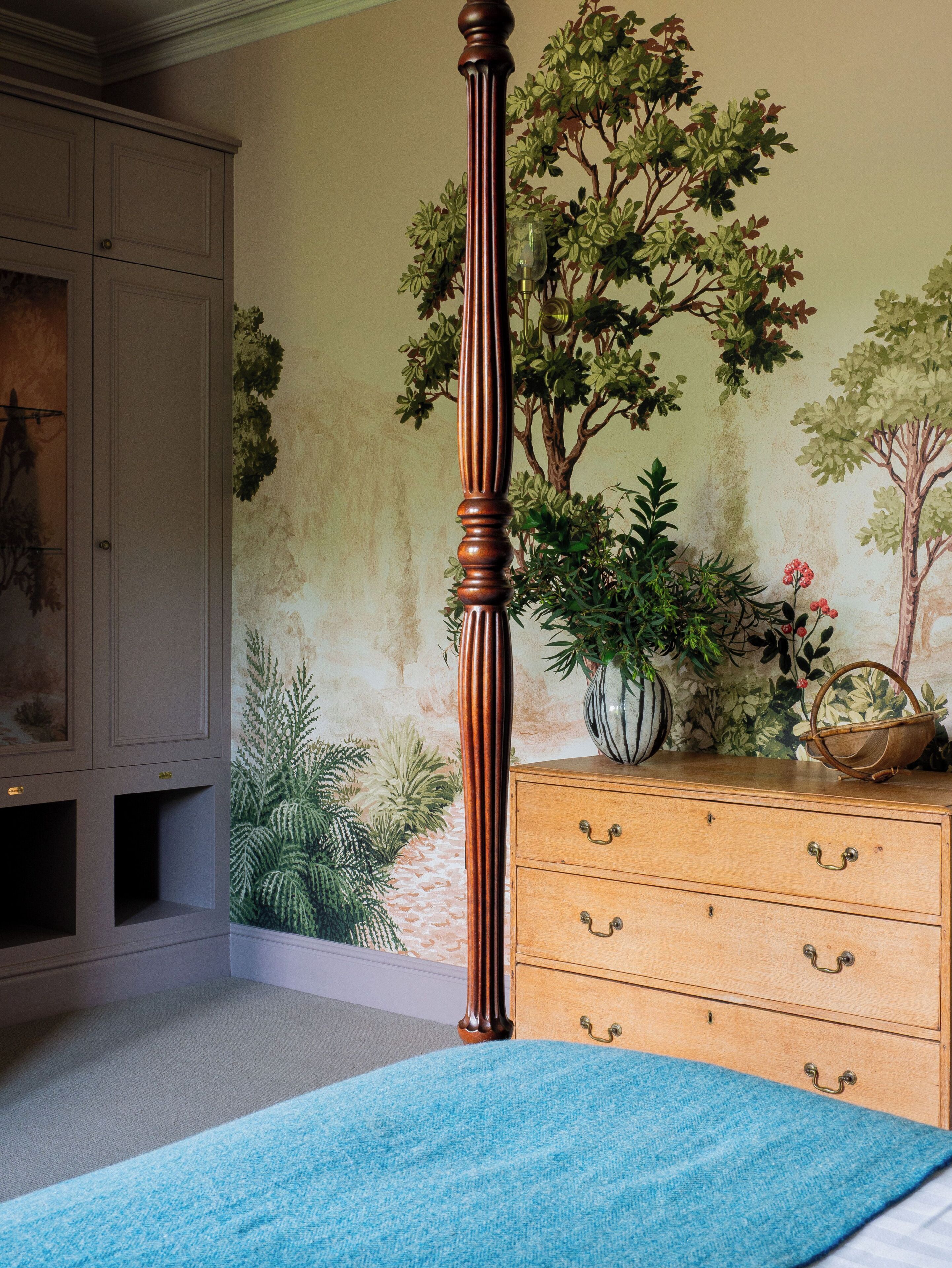 A bedroom with a four-poster bed, a wooden dresser, and a wall mural of trees.