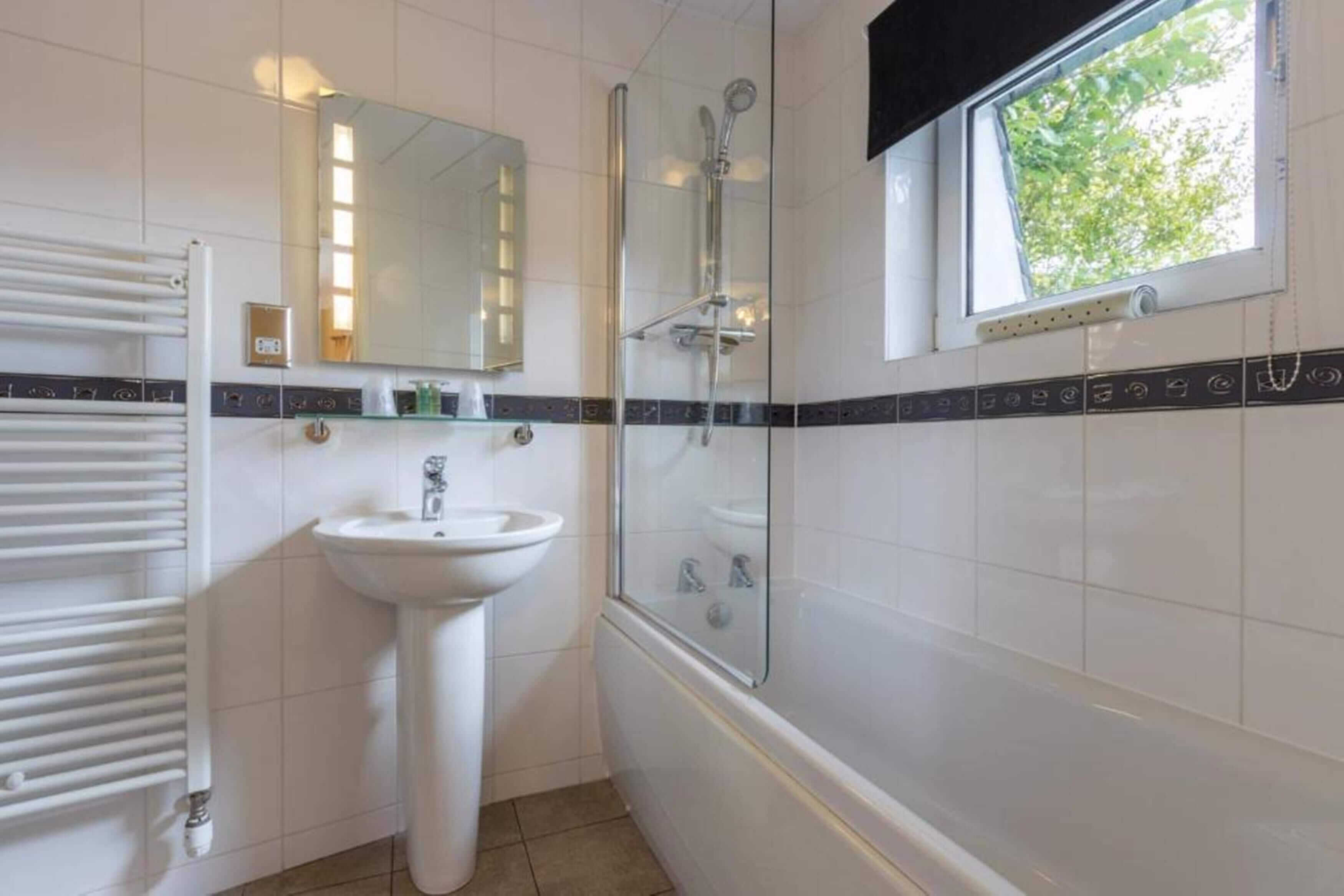 Un baño con un lavamanos blanco, ducha con mampara de vidrio, toallero y ventana con vista a la vegetación.
