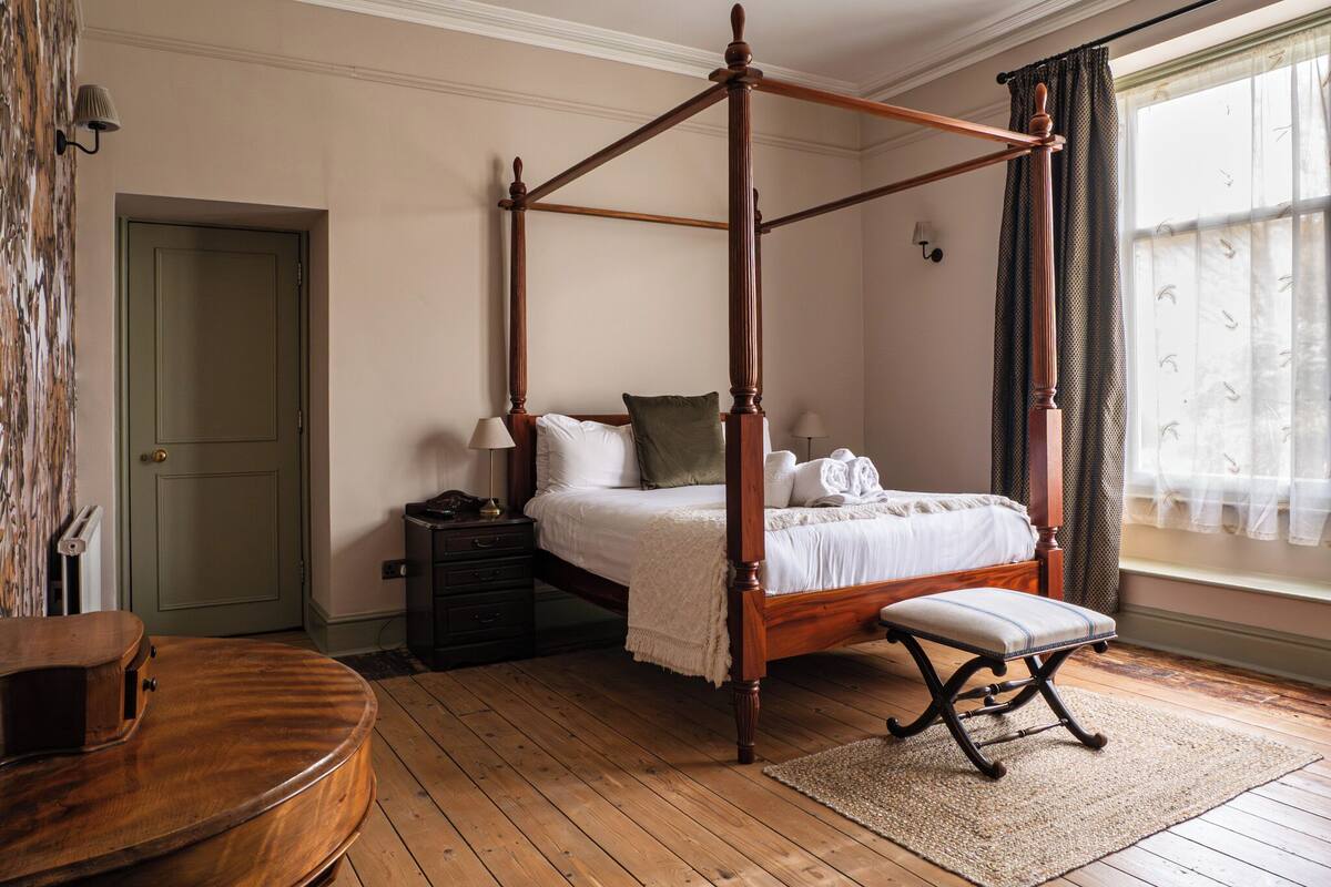 A four-poster bed with a wooden finish, a bedside table with a lamp, a bench at the foot of the bed, and a window with curtains.