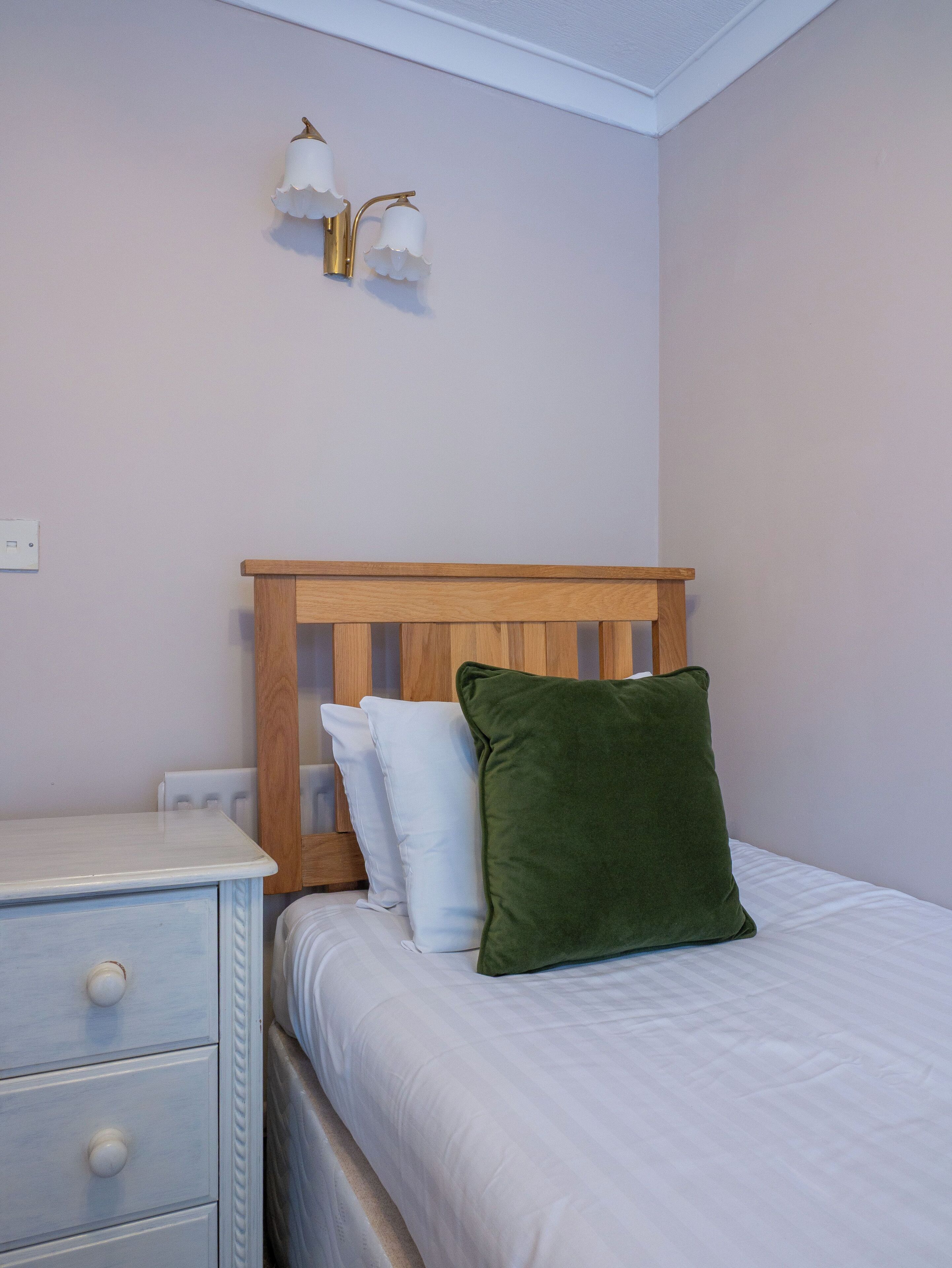 Un dormitorio con cabecera de madera, una mesita de noche blanca, un almohadón verde y una lámpara de pared.