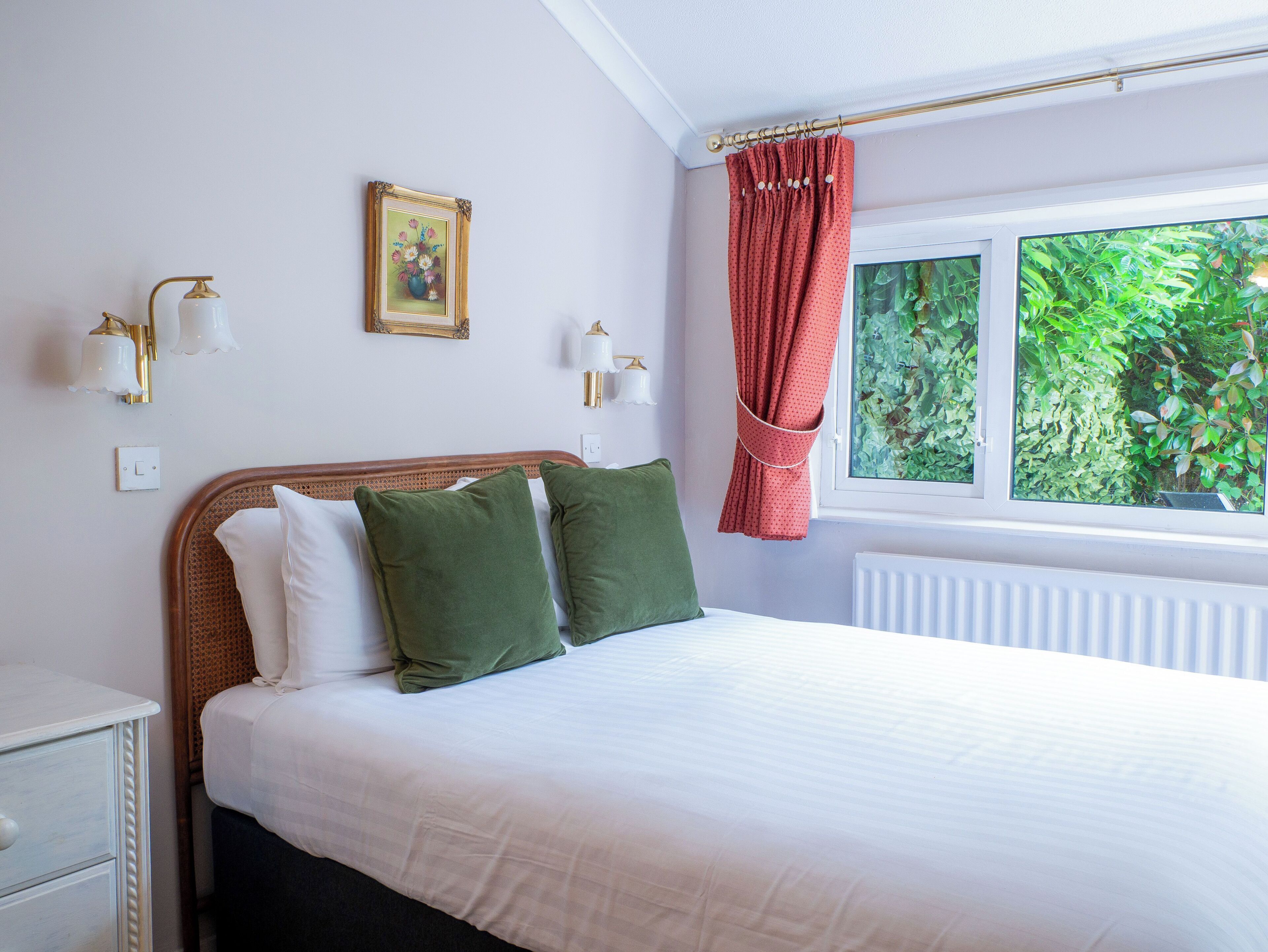 A neatly made bed with white and green pillows, a framed picture on the wall, and a window with a view of greenery.