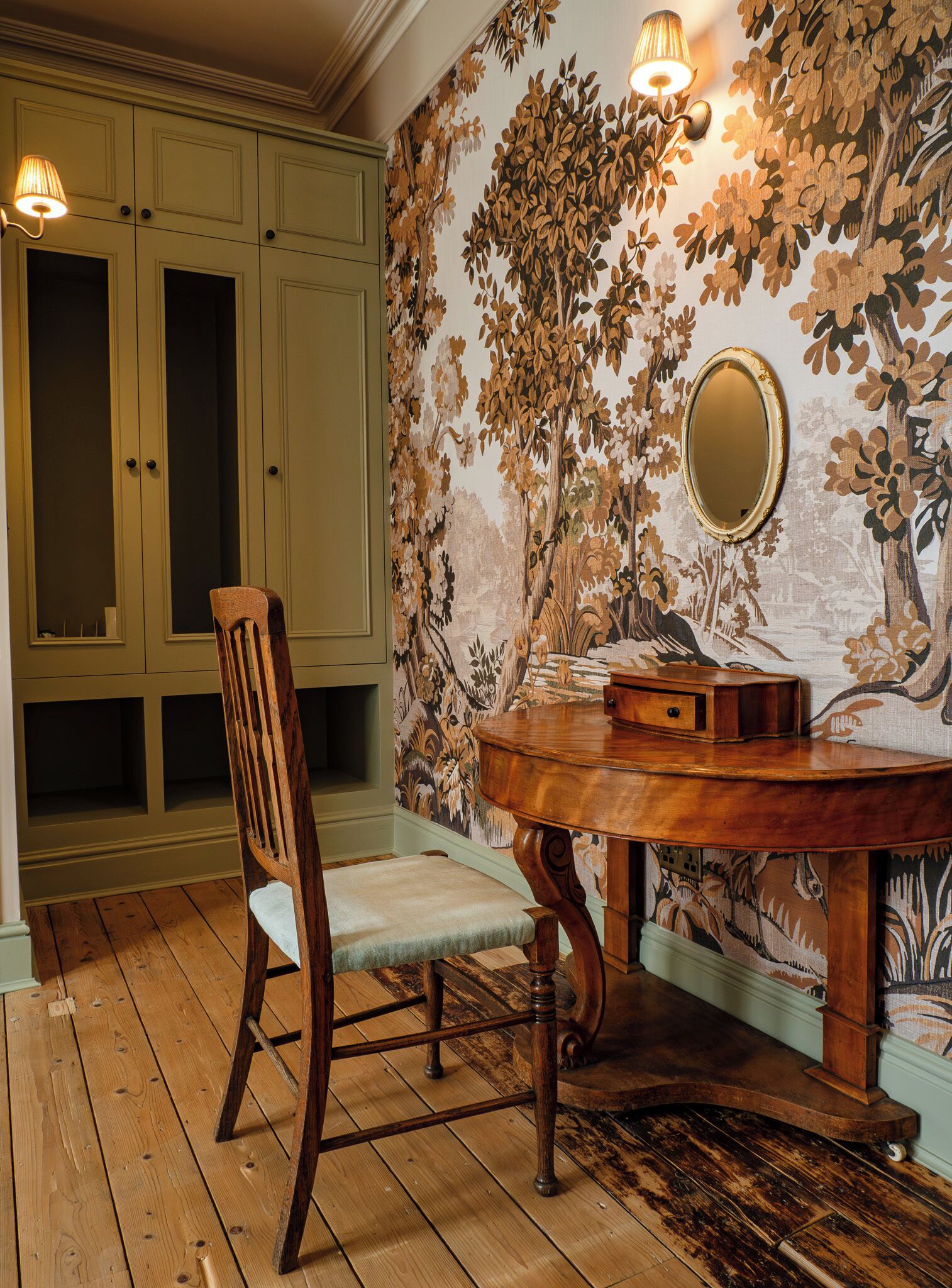 A room with a wooden chair, a small table, and a wall with floral wallpaper. There are two cabinets with doors and a light fixture above the table.