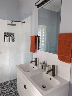 Une salle de bain avec un lavabo blanc, deux robinets, un miroir et une pomme de douche. Deux serviettes sont accrochées au mur.