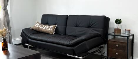 A modern living room with a black leather sofa, a small wooden side table, a glass coffee table, and a black and white rug.