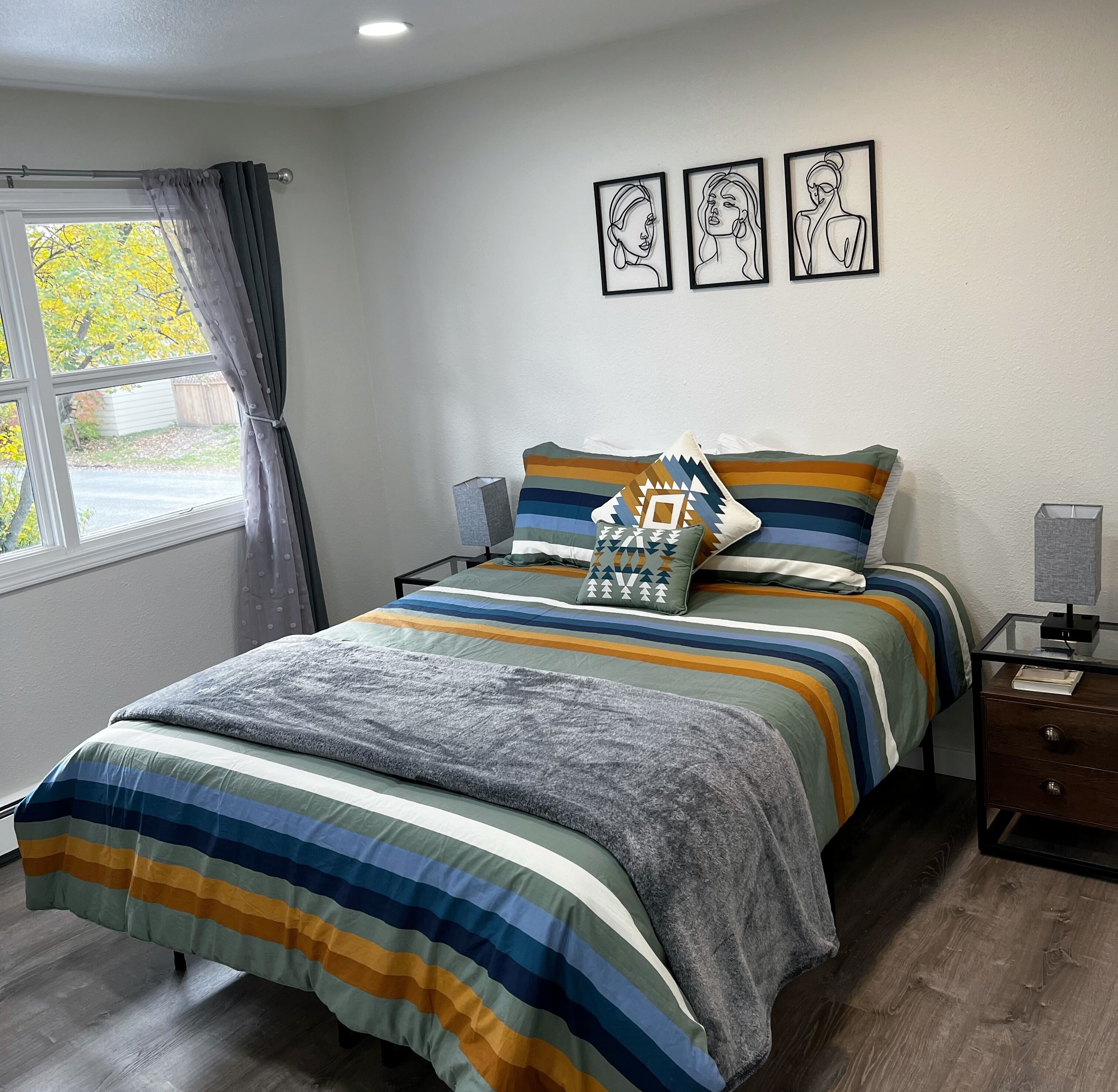 Un dormitorio con cama, mesita de noche, lámpara y ventana con cortinas. Hay tres cuadros en la pared.