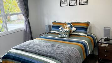 Un dormitorio con cama, mesita de noche, lámpara y ventana con cortinas. Hay tres cuadros en la pared.