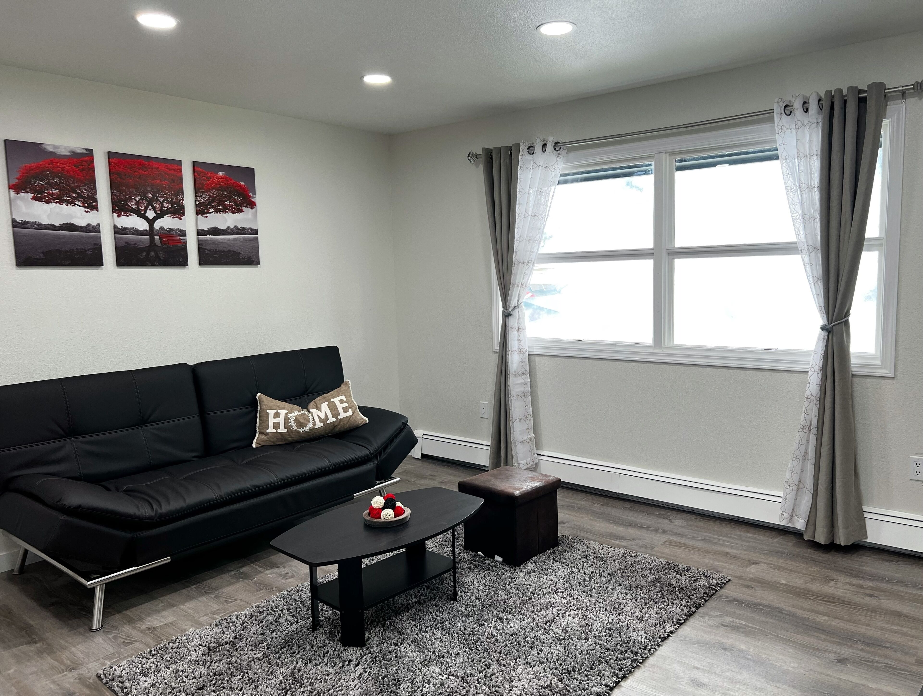 A modern living room with a black leather sofa, a coffee table, and a small ottoman. There are three framed pictures on the wall, a window with curtains, and a rug on the floor.