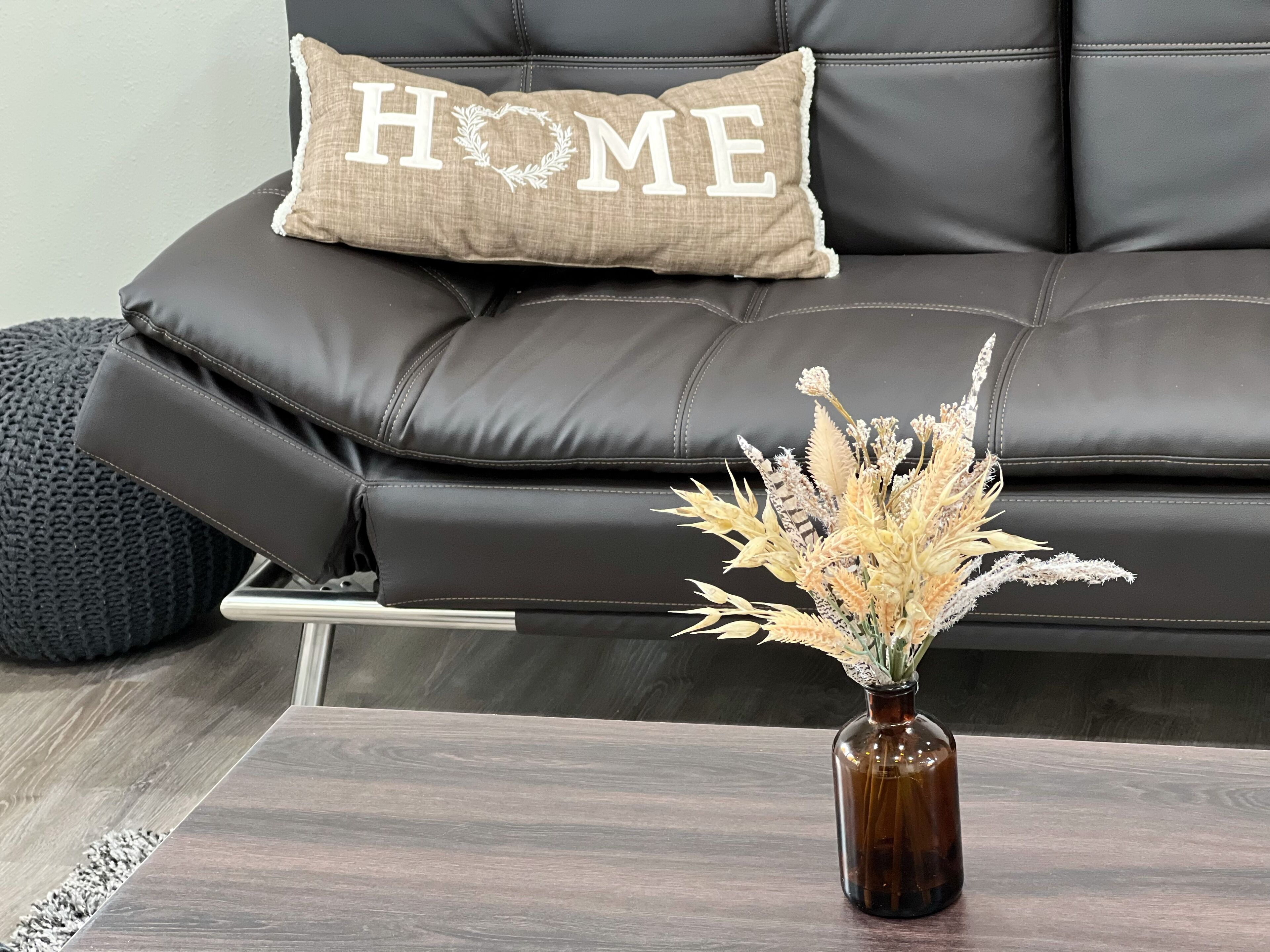A brown leather sofa with a cushion that has "HOME" embroidered on it. In front of the sofa, there is a small brown vase with dried flowers.