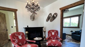 Un salón con chimenea, dos sillones rojos con estampado de ciervo, una araña y una cabeza de ciervo montada en la pared.