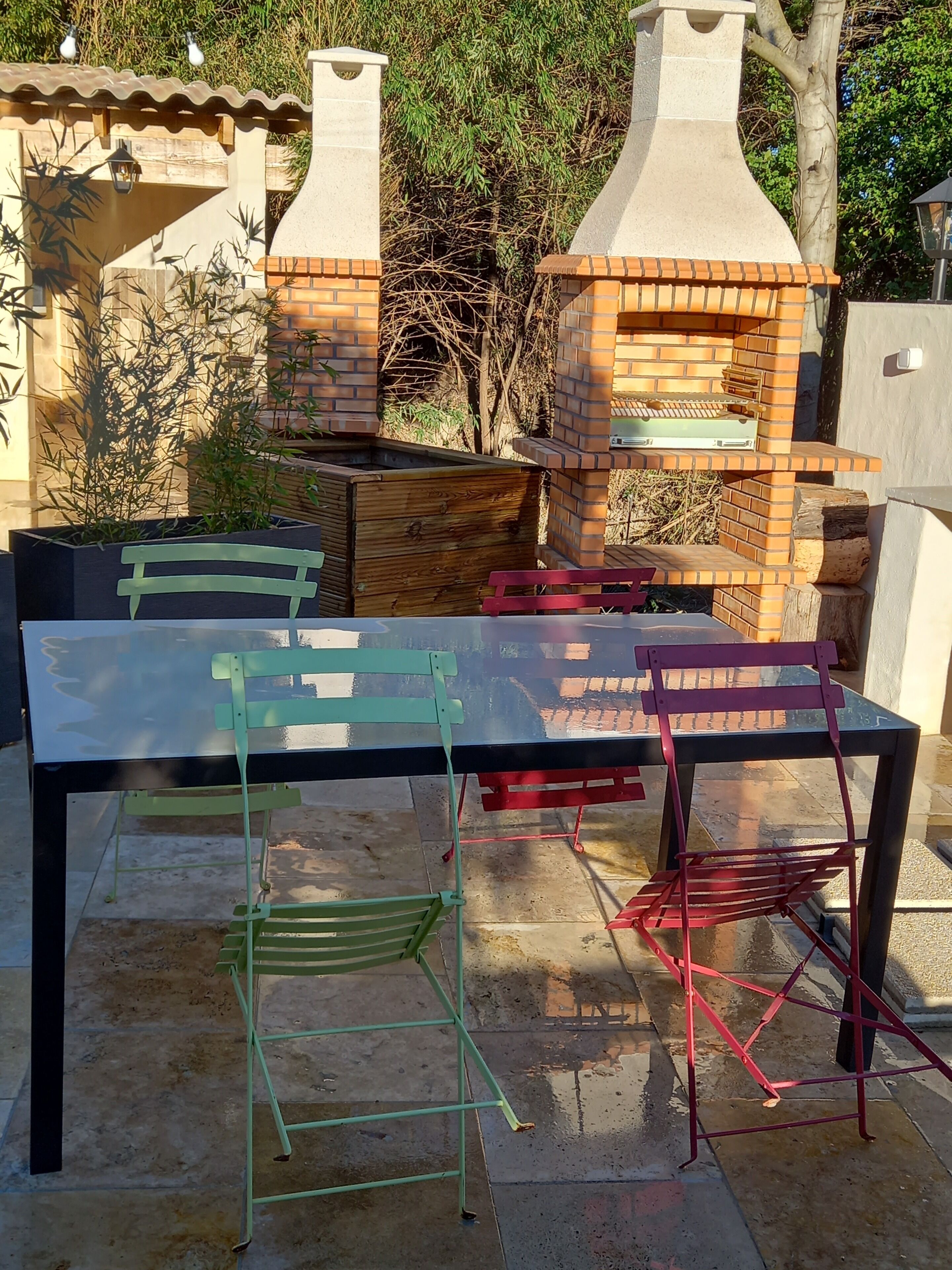 Une terrasse extérieure avec une table en verre, deux fours en briques et des chaises pliantes.