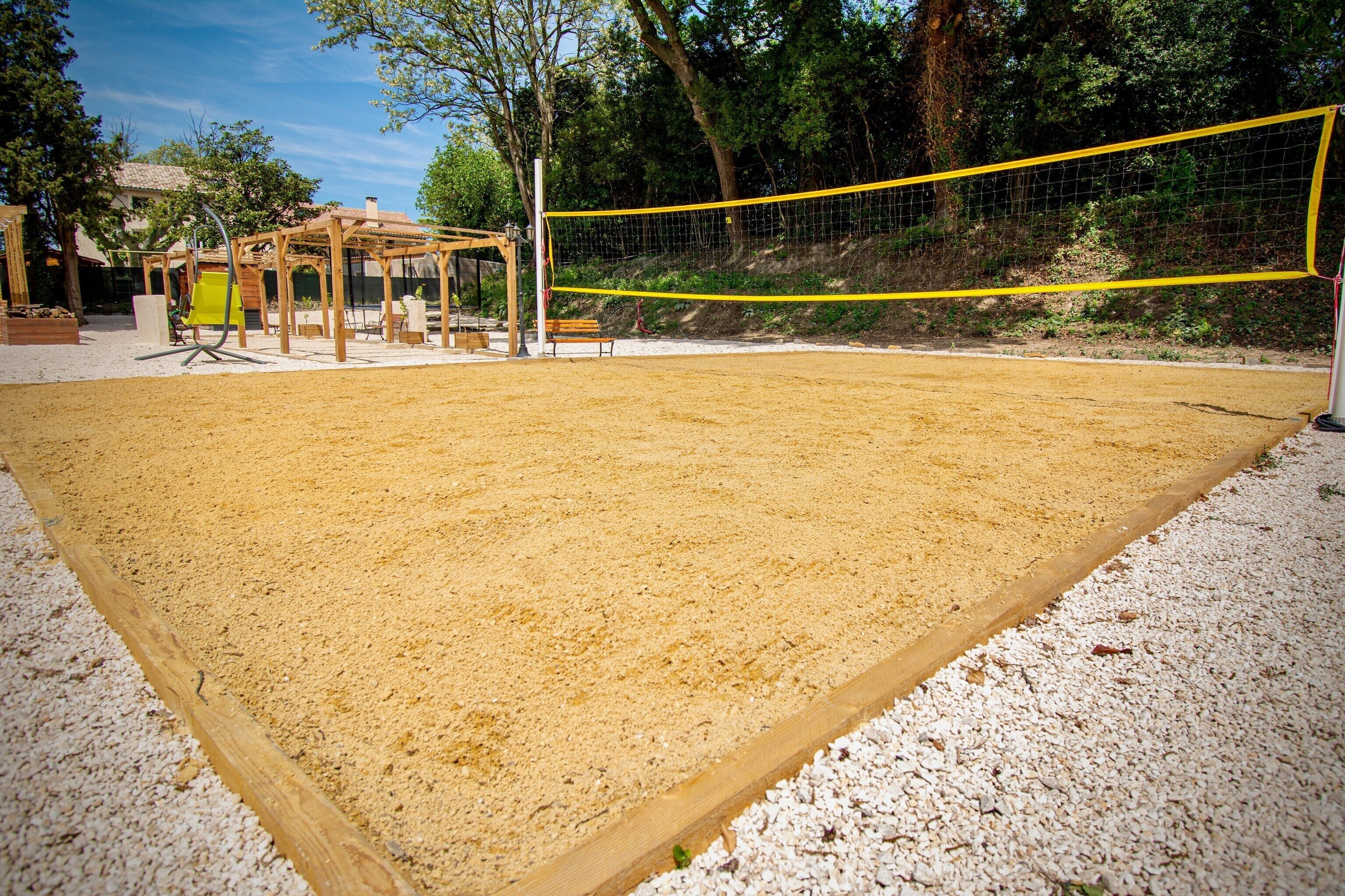 Una cancha de voleibol al aire libre con superficie de arena, rodeada de árboles y una estructura de pérgola de madera.