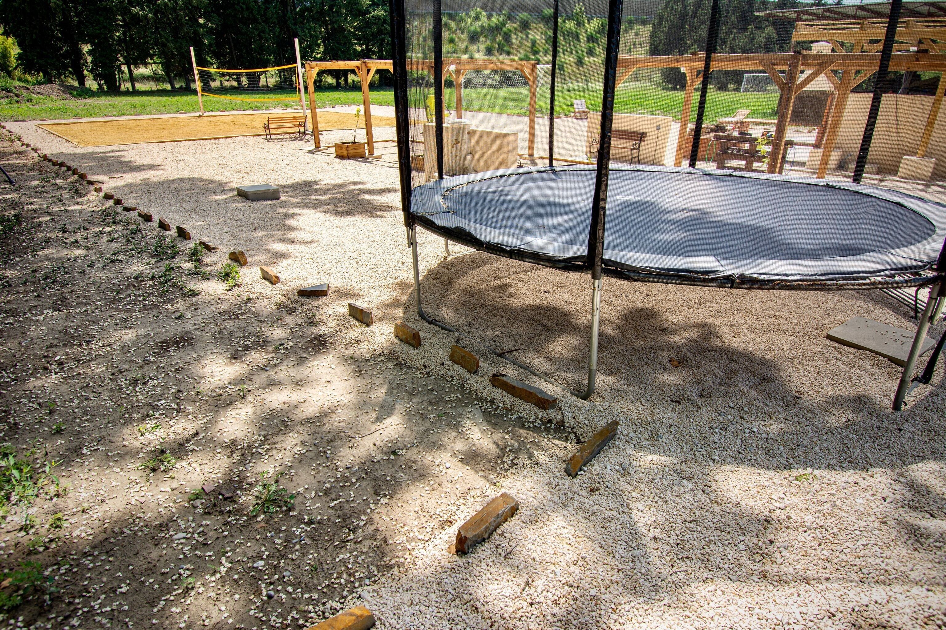 A trampoline surrounded by a wooden fence, with a gravel path leading to a wooden deck.