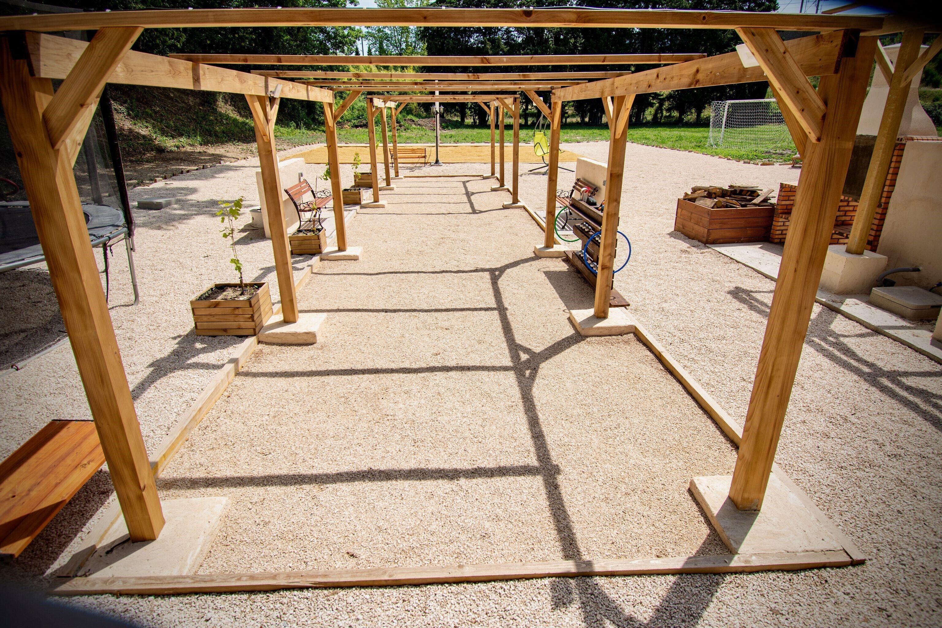 A wooden pergola structure with a gravel floor, surrounded by potted plants and wooden benches.
