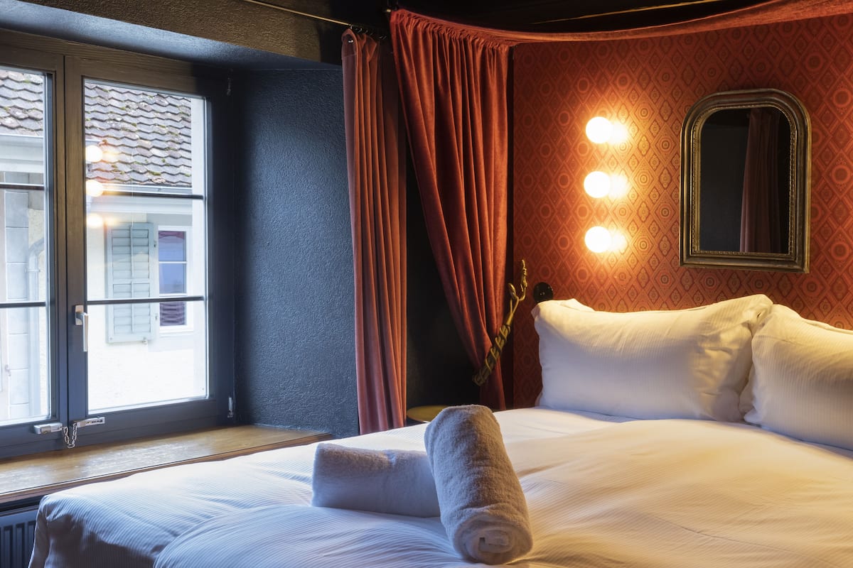Quarto de hotel com uma cama grande, uma janela com cortinas, um espelho e luminárias fixadas na parede.