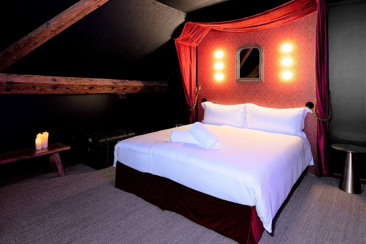 Quarto com teto de madeira, cama com roupa de cama branca, mesa de cabeceira pequena e cortina vermelha.