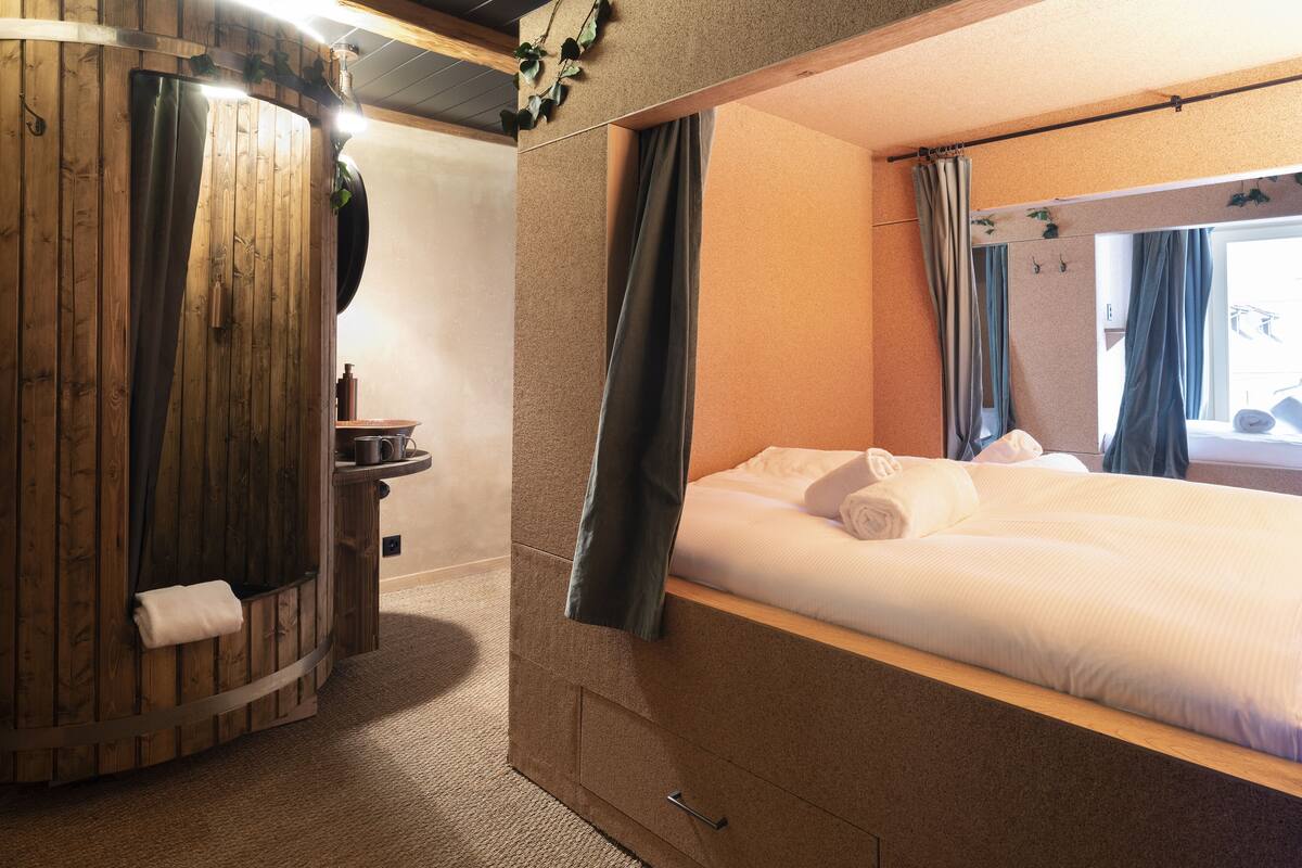 Quarto de hotel compacto, com cama, banheiro pequeno e janela com cortinas.