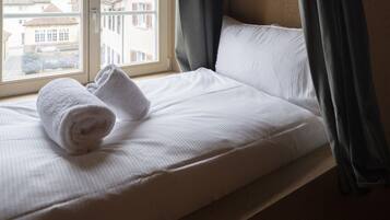Un lit bien fait, avec du linge de lit blanc et des serviettes pliées, une fenêtre avec des rideaux et une vue sur les bâtiments à l’extérieur.