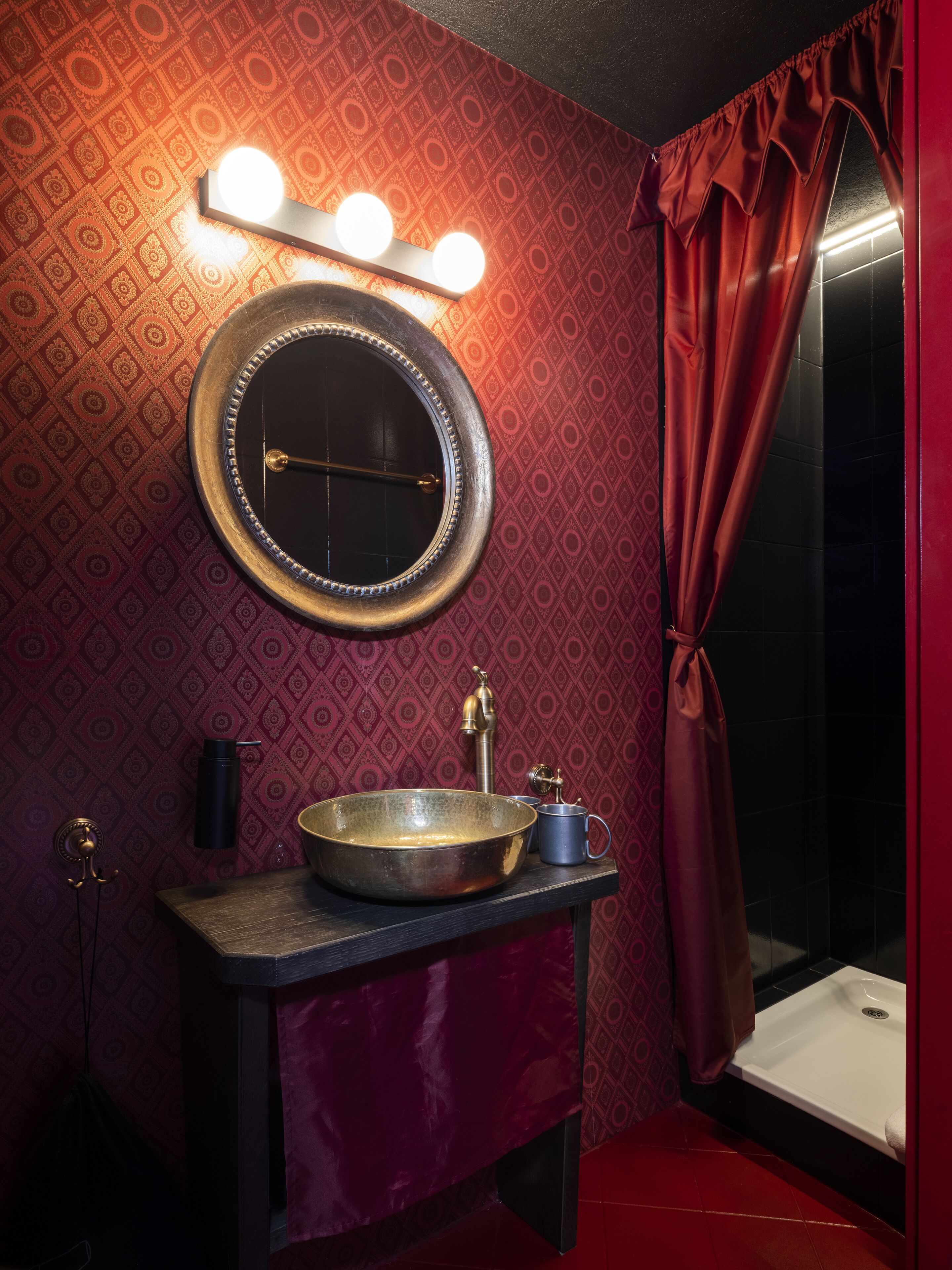 Un baño con espejo redondo, tocador, lavamanos y cortinas rojas.