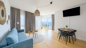 A modern living room with a blue sofa, a dining table with chairs, a TV mounted on the wall, and a view of the sea through large windows. - Apartamenty Sun&Snow Przy Plaży (Mielno)