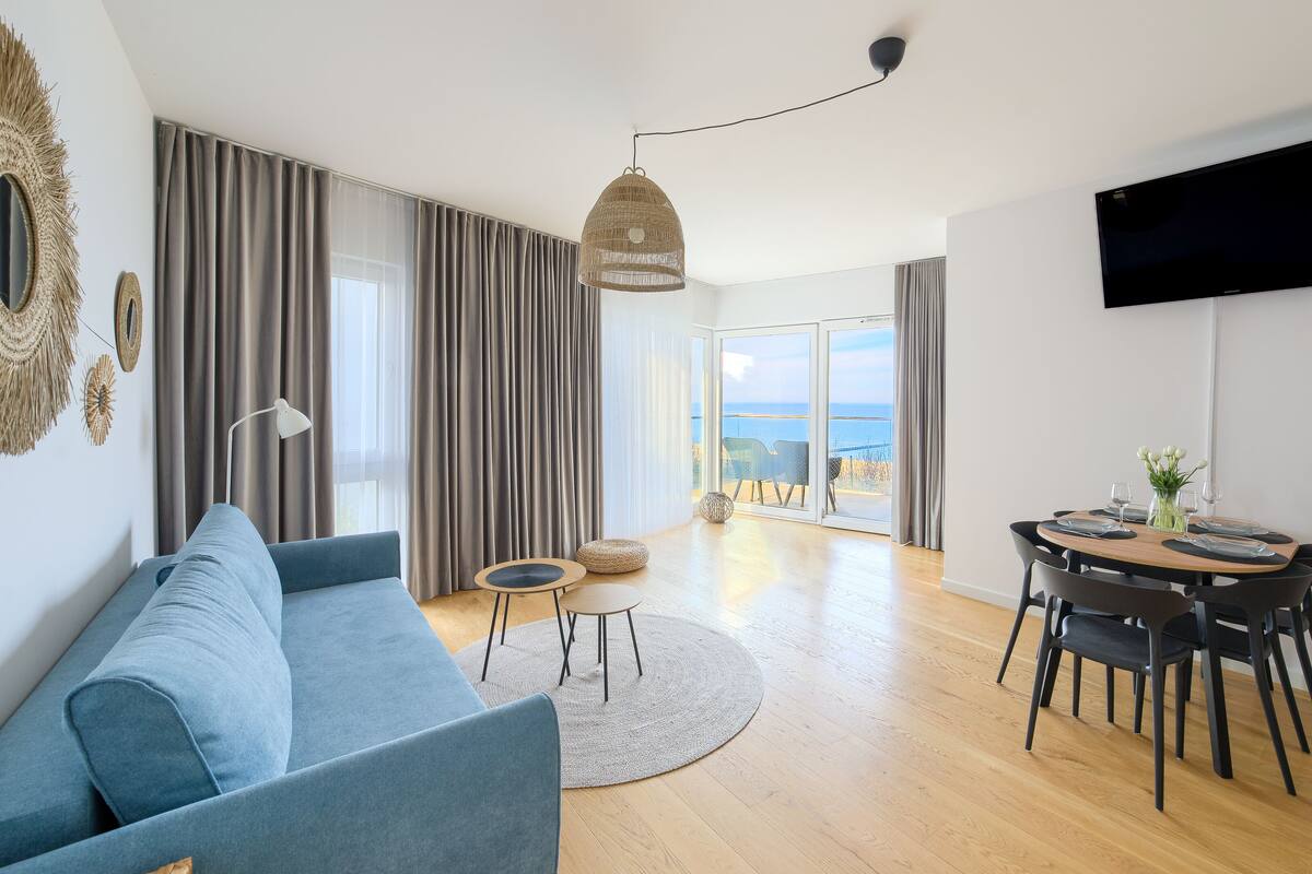 A modern living room with a blue sofa, a dining table with chairs, a TV mounted on the wall, and a view of the sea through large windows.