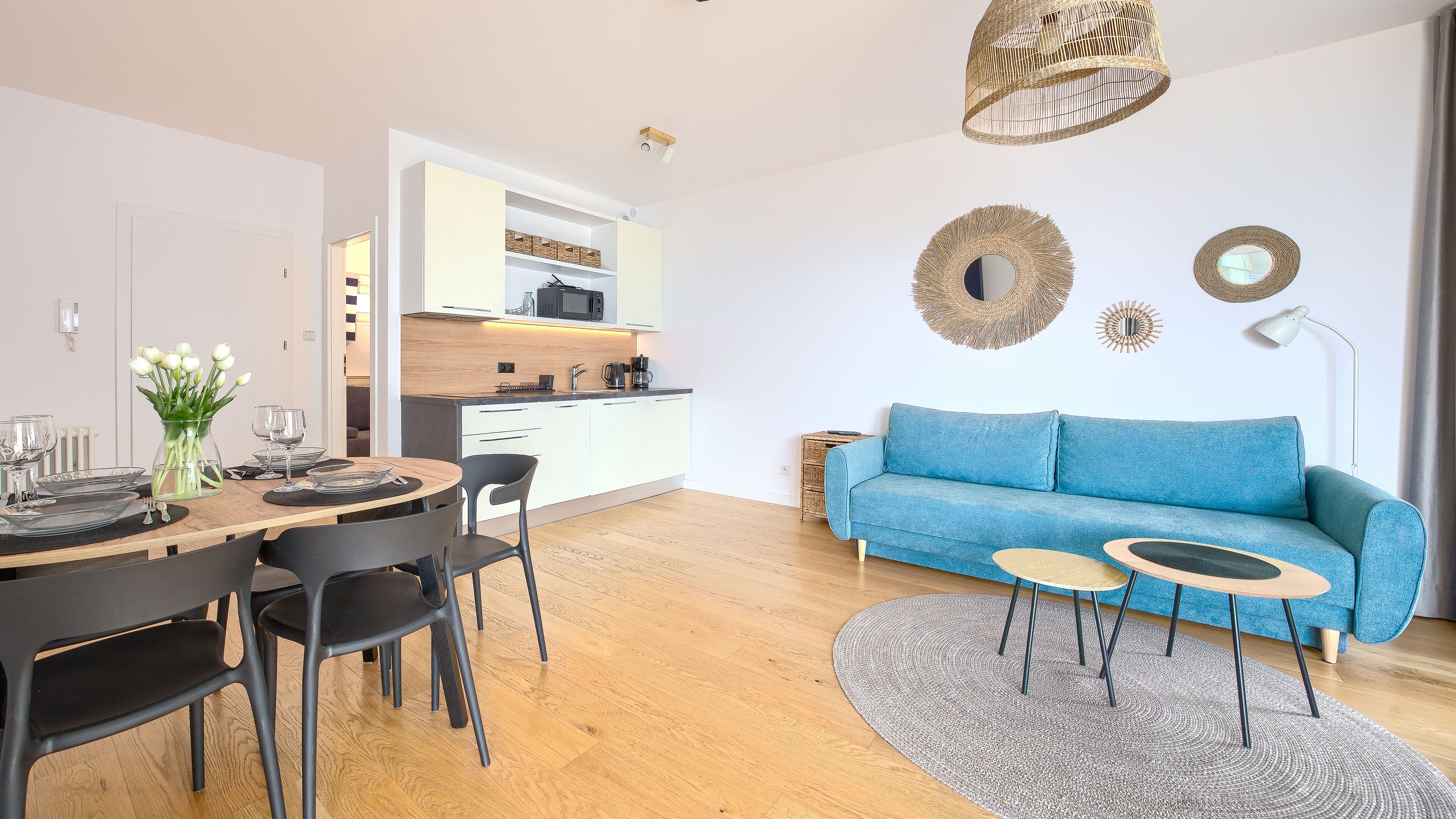 Una sala moderna con un sofá azul, una mesa de comedor dispuesta con platos y vasos, un jarrón de flores y una cocina al fondo.