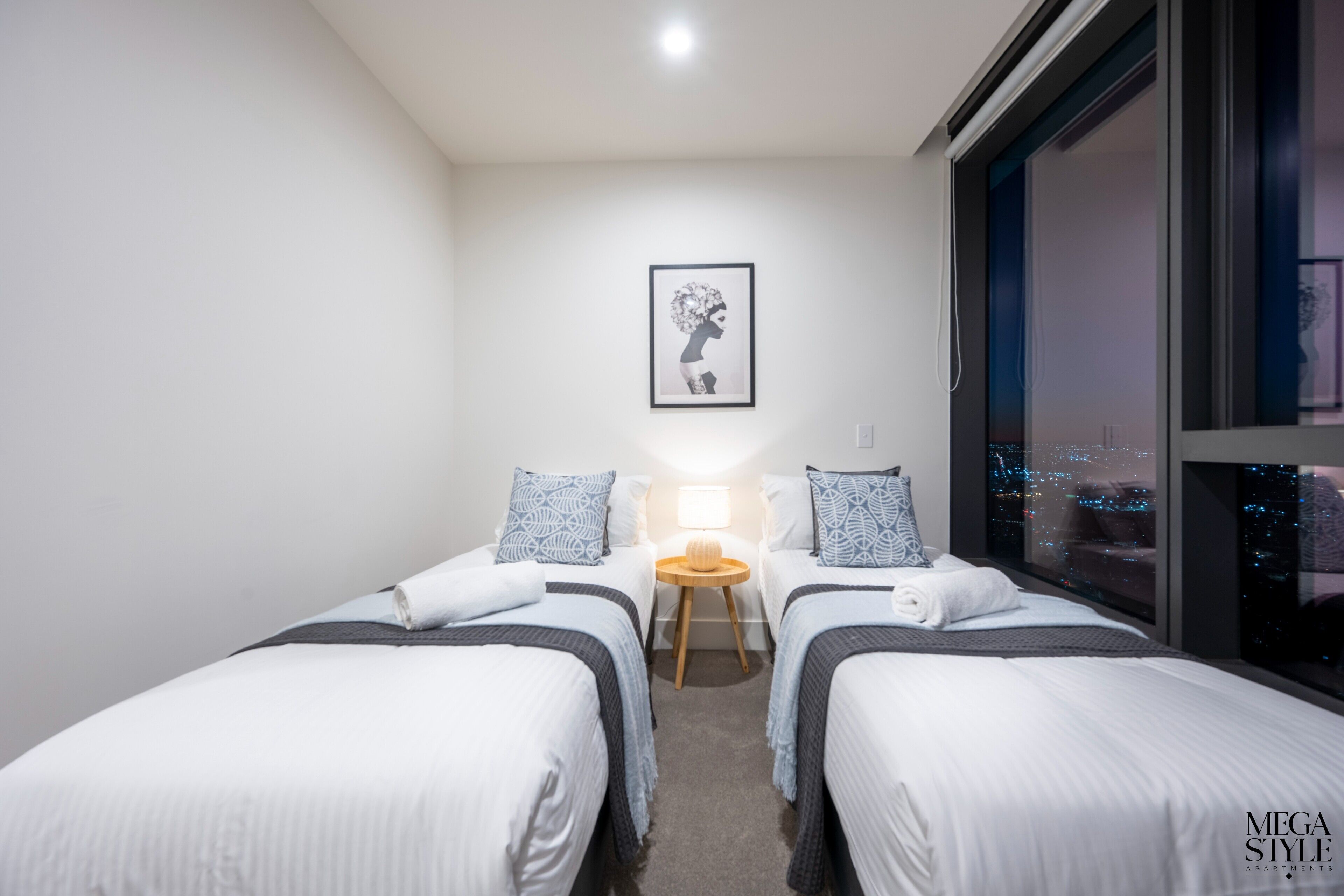 A modern hotel room with two single beds, a nightstand with a lamp, a framed artwork on the wall, and a large window offering a city view.