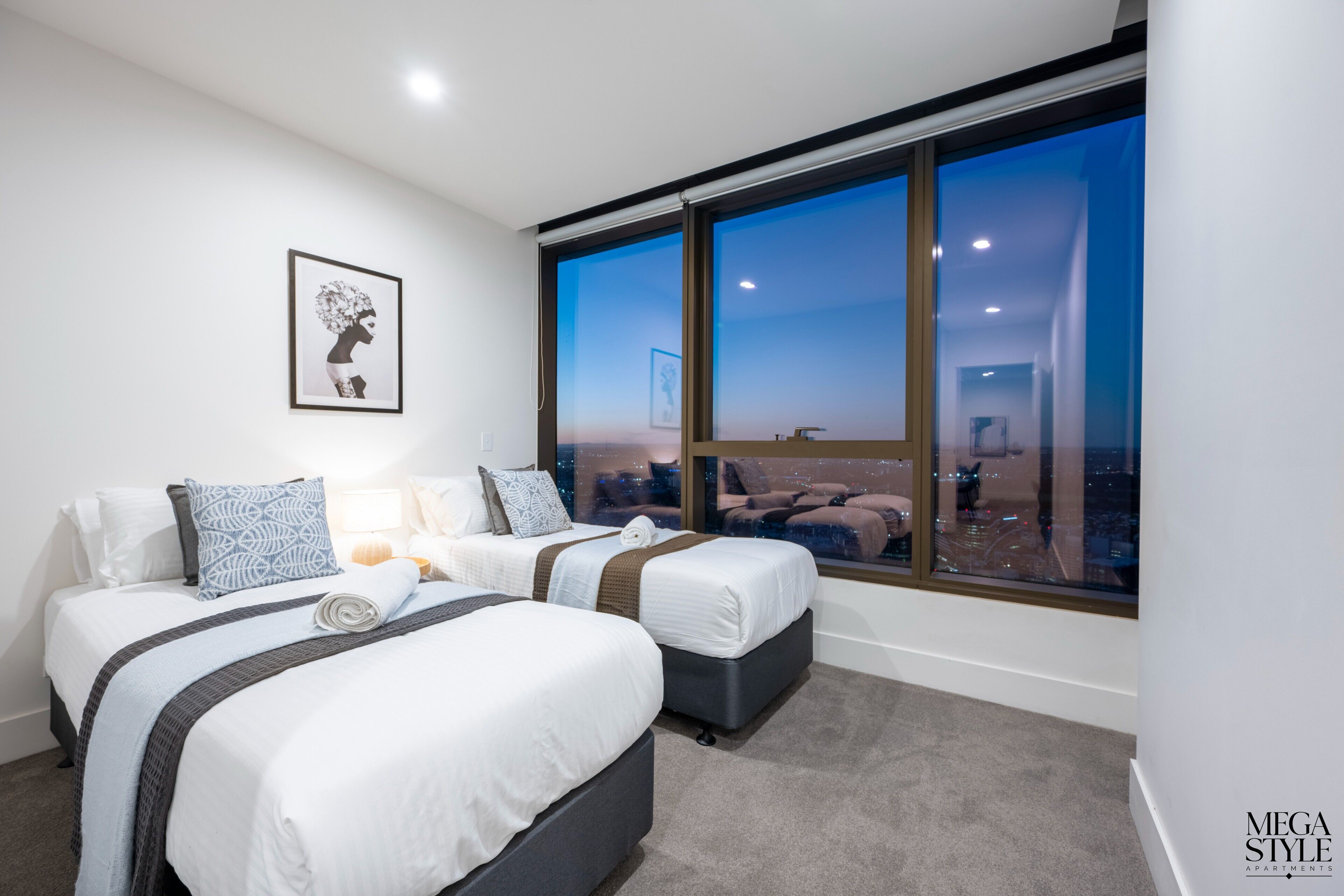 Ein modernes Schlafzimmer mit zwei Betten, einem großen Fenster mit Blick auf die Stadt und einem gerahmten Kunstwerk an der Wand.