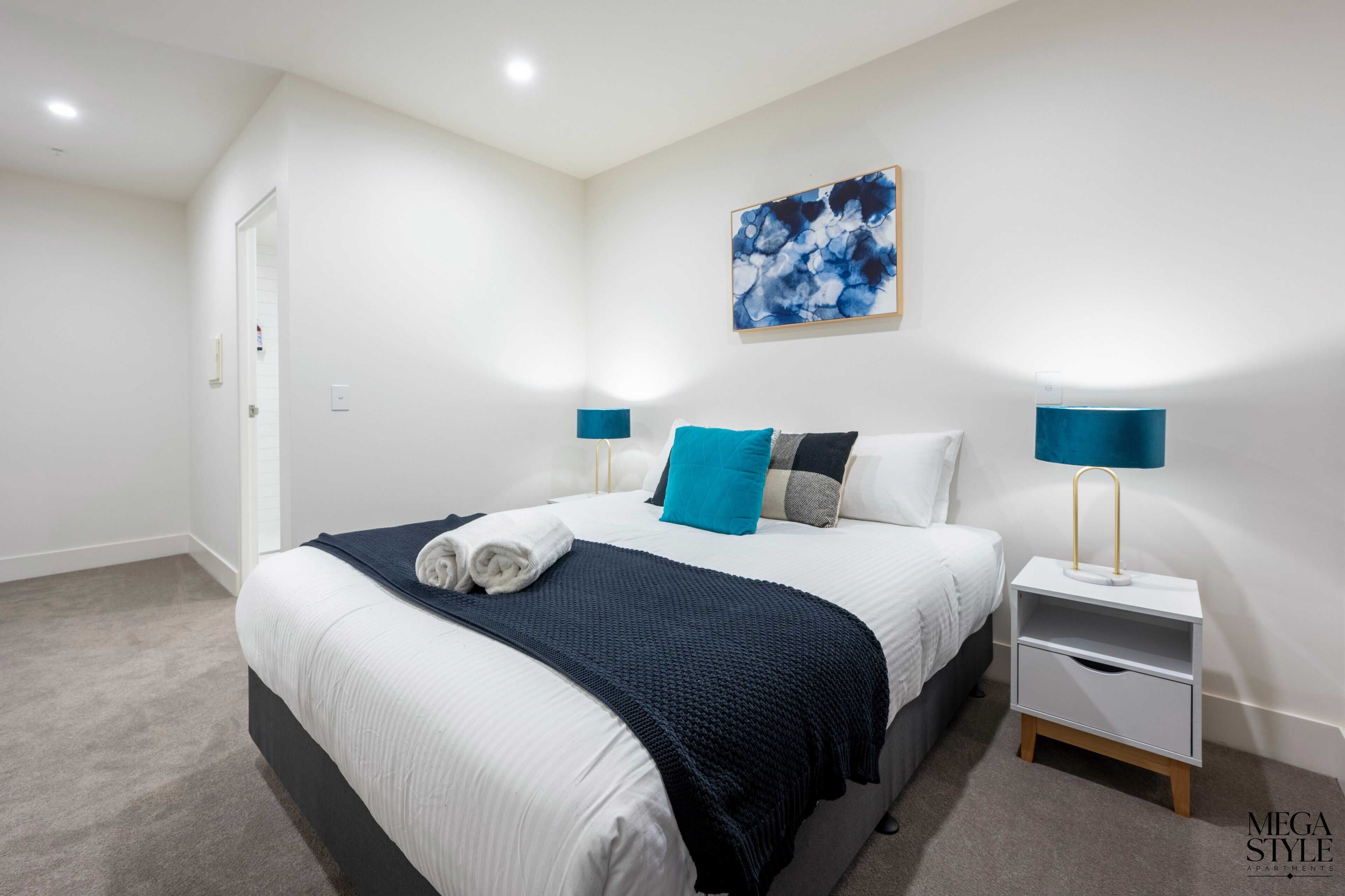 Ein modernes Schlafzimmer mit einem ordentlich bezogenen Bett, einem blauen abstrakten Gemälde und einem Nachttisch mit Lampe.